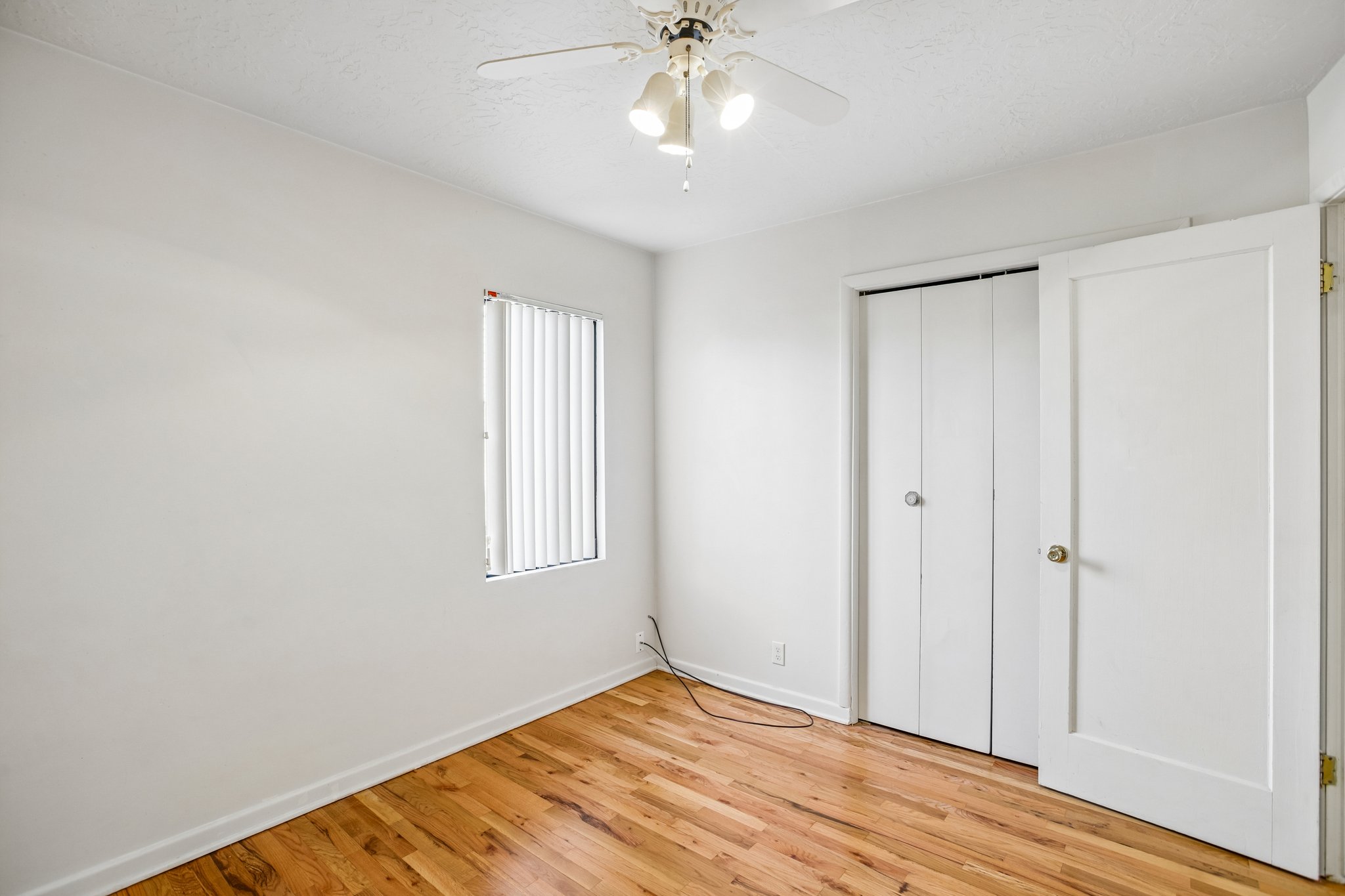 Bright room featuring wood-finish flooring, white walls, a window with vertical blinds, a ceiling fan with integrated lighting, and a closet with bifold doors