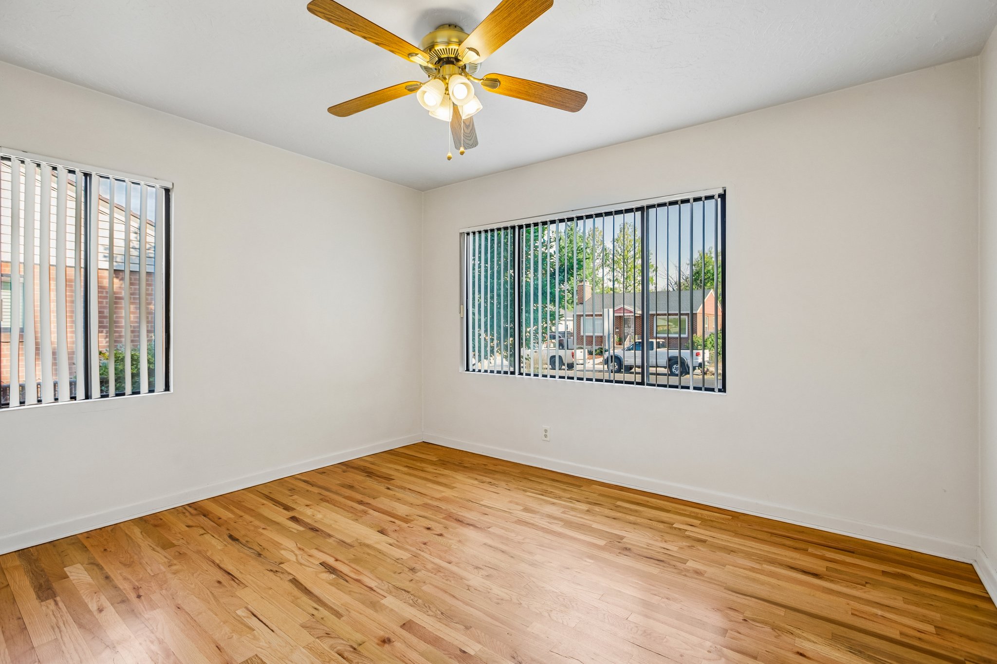 Spacious room featuring wood-finish flooring, two windows with vertical blinds, a ceiling fan with integrated lighting, and white walls