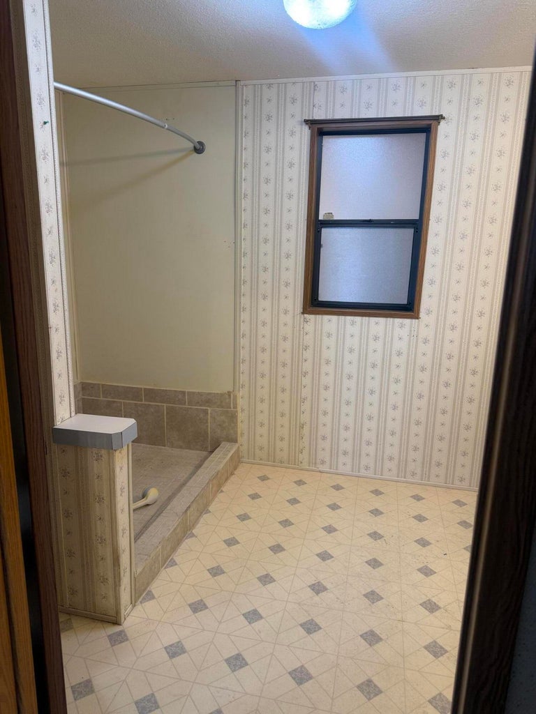Bathroom featuring a built-in shower enclosure with a tiled base and walls, a frosted window with dark wood trim, patterned wallpaper, and geometric pattern flooring