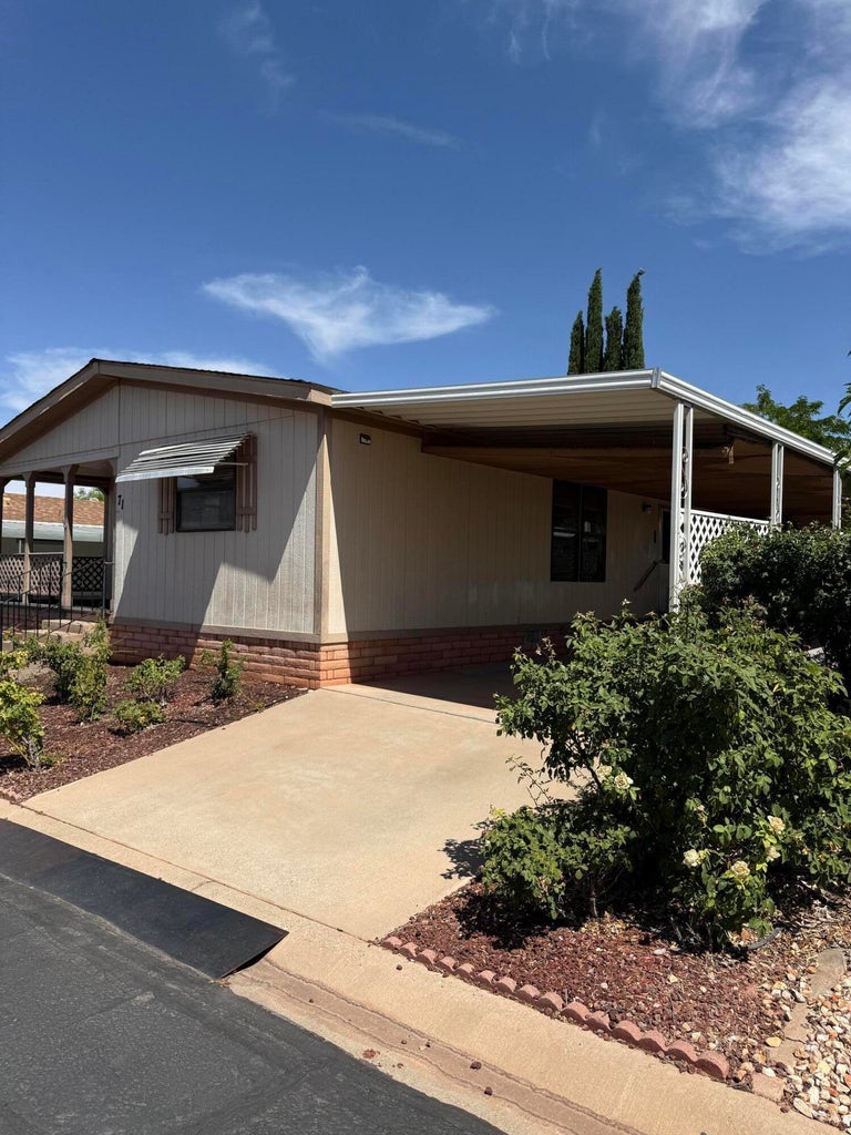 Exterior featuring a covered carport, mature landscaping, paved driveway, and brick-look skirting
