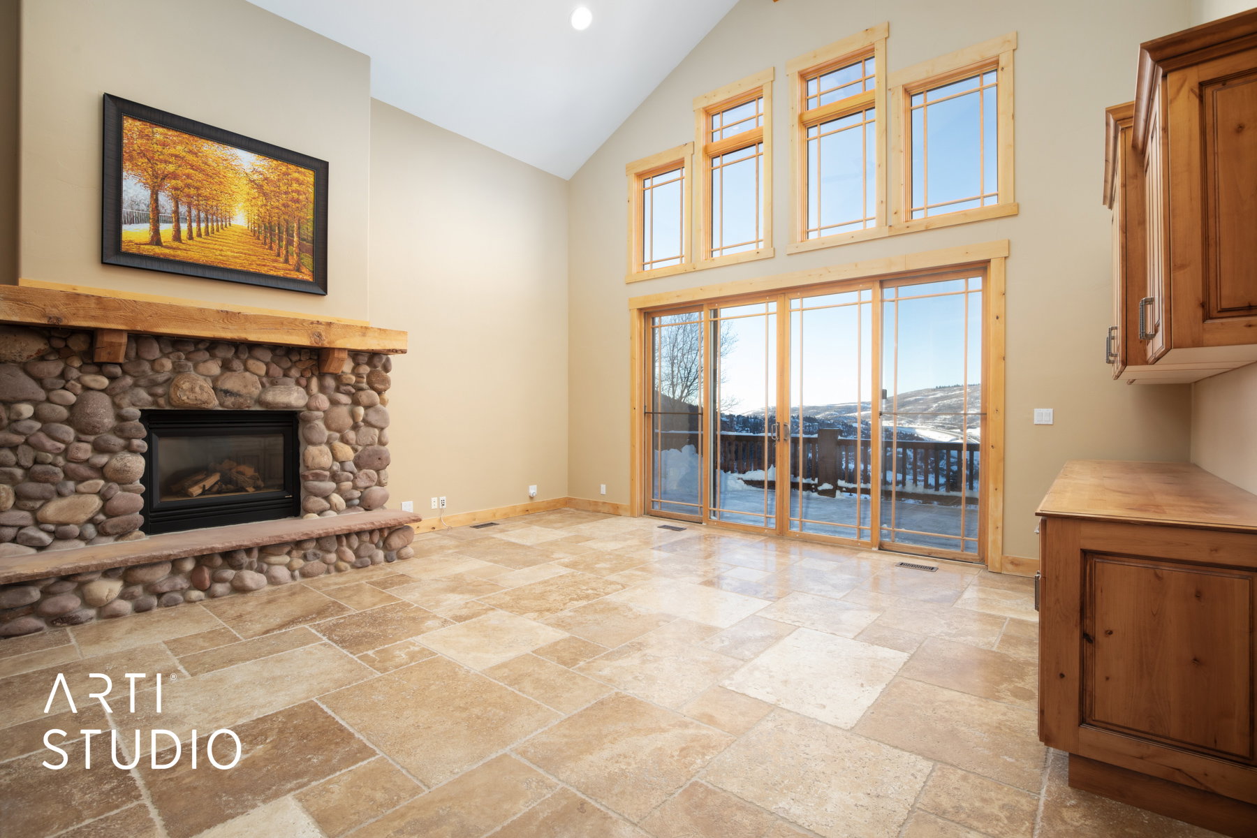 Vaulted ceiling living area featuring a stacked stone fireplace with a substantial timber mantel, large format tile flooring, and a wall of windows and sliding glass doors opening to a deck