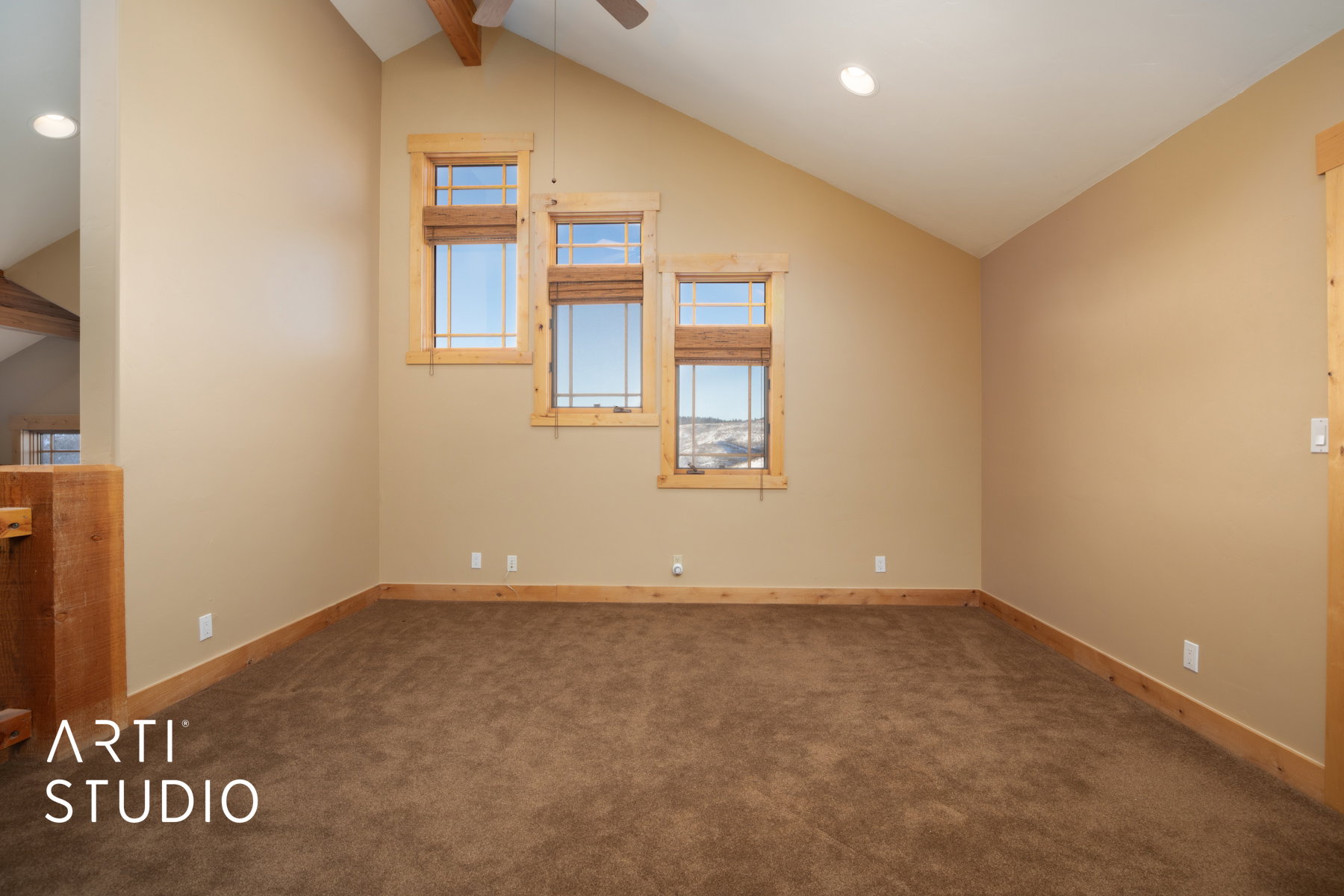 Vaulted ceiling room featuring three staggered windows with natural wood trim, coordinating wood baseboards, and plush brown carpeting