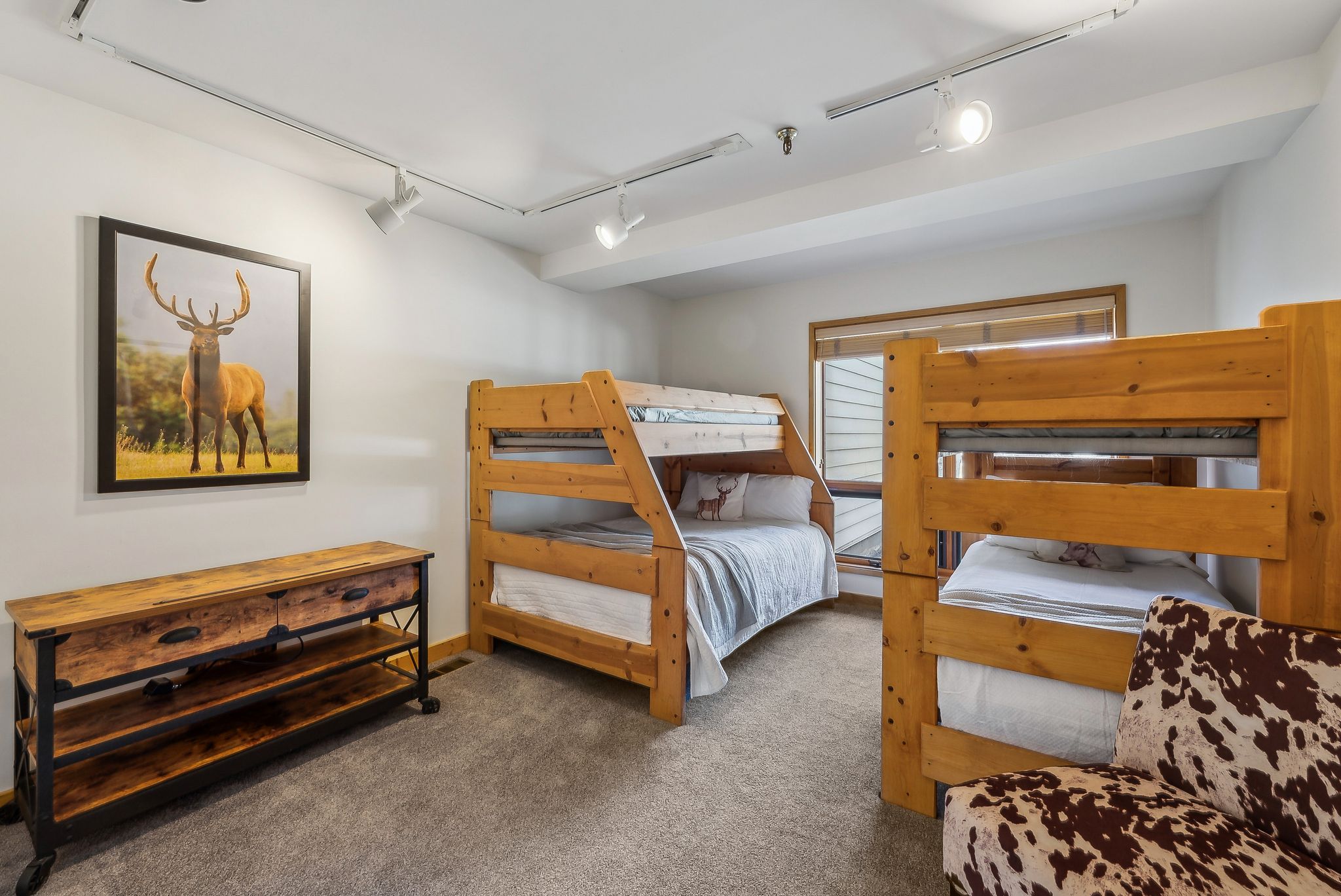 Carpeted room featuring two wooden bunk bed structures, a window with wood trim, track lighting, and a rustic wood-finish console table with metal accents