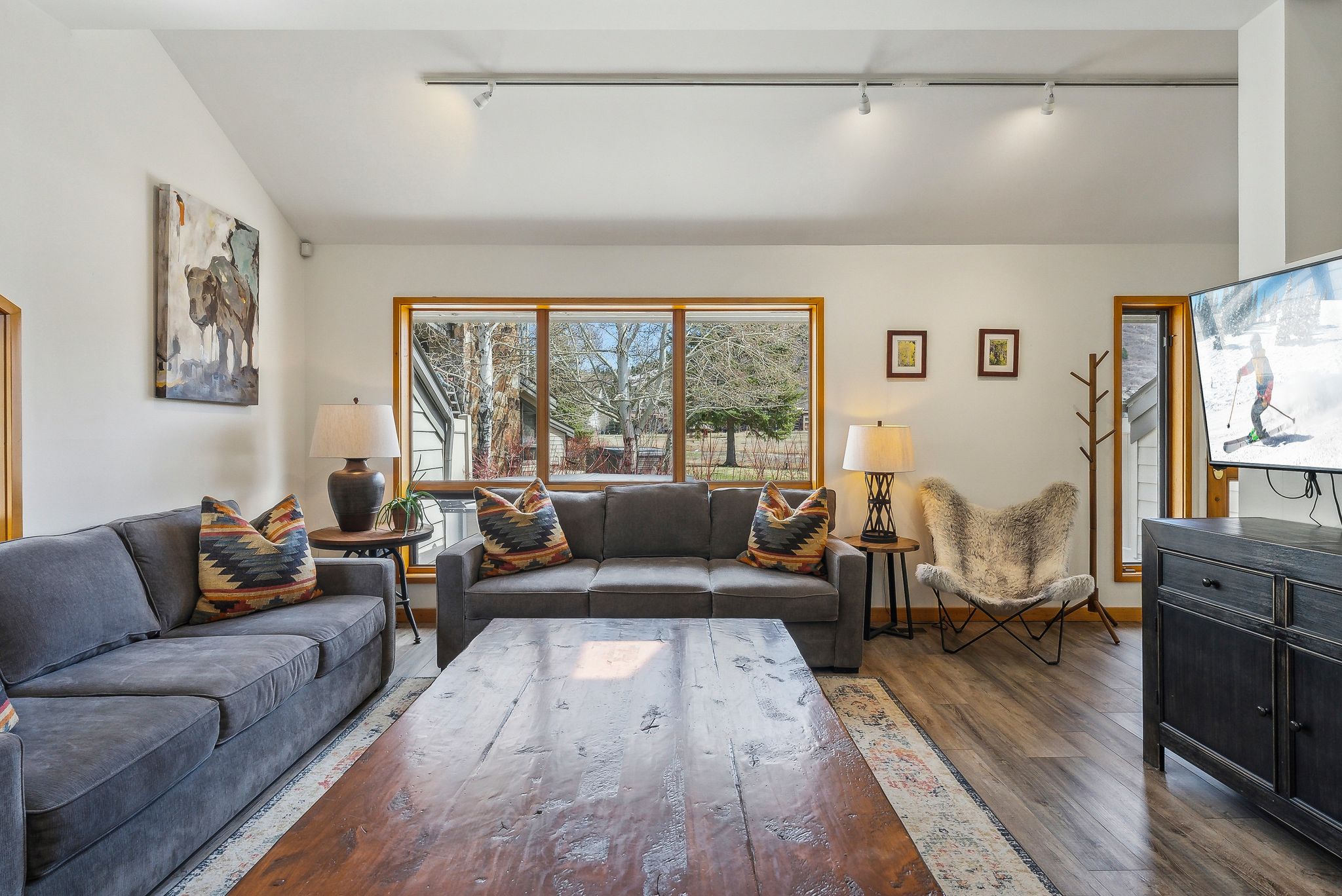 Spacious living area featuring vaulted ceilings, track lighting, wood-finish flooring, and large windows with natural wood trim