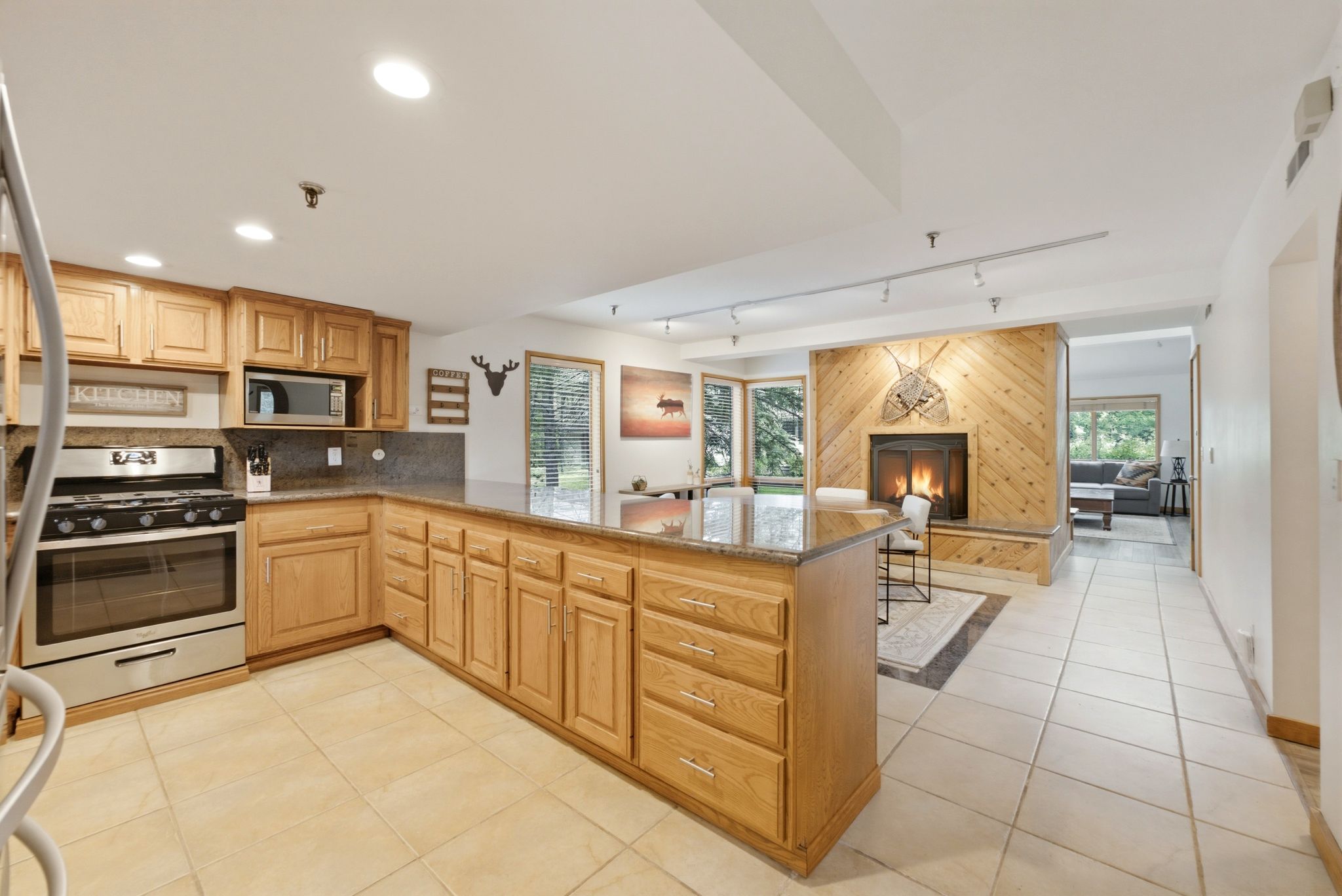 Open-concept kitchen and living area featuring a stone countertop island, stainless steel appliances, abundant cabinetry, and a wood-paneled fireplace
