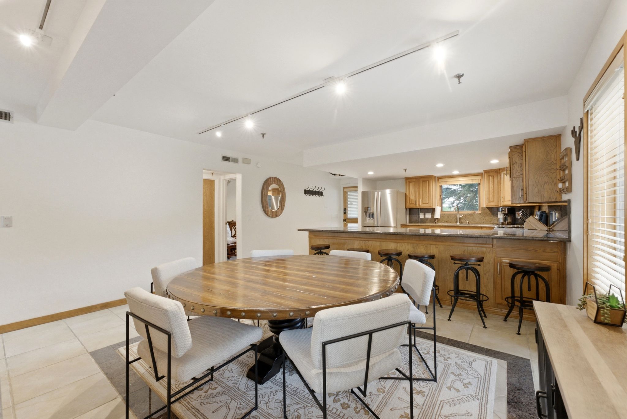 Open-concept living area featuring a wood-finish dining table with stud detailing, track lighting, light tile flooring, and a kitchen with wood cabinetry and stainless steel appliances