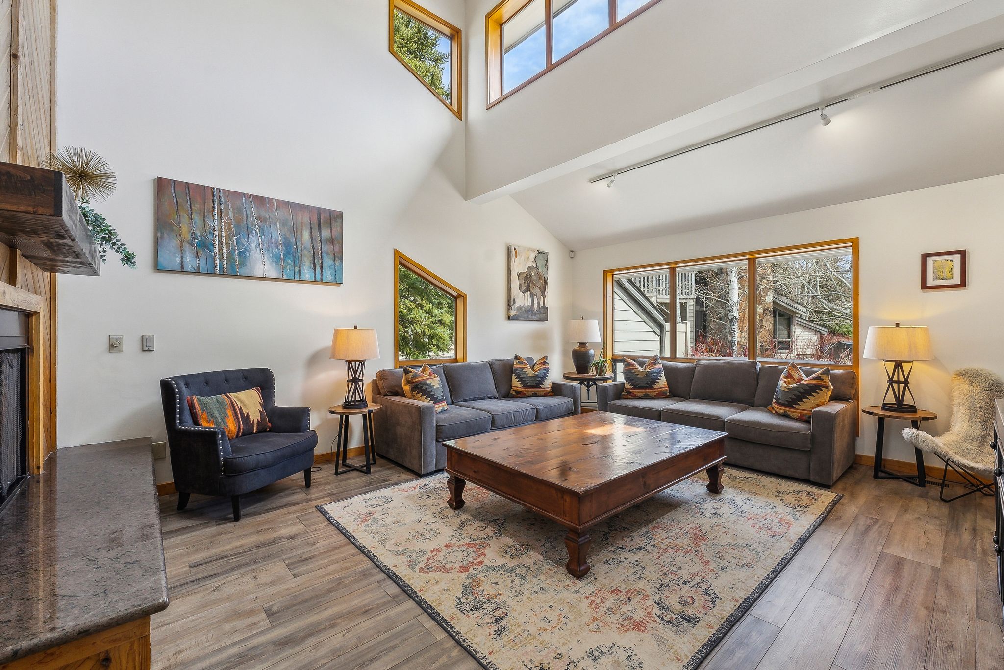 Vaulted ceiling living room featuring wood-finish flooring, a stone-topped built-in media console with a fireplace insert, and multiple large windows with natural wood trim