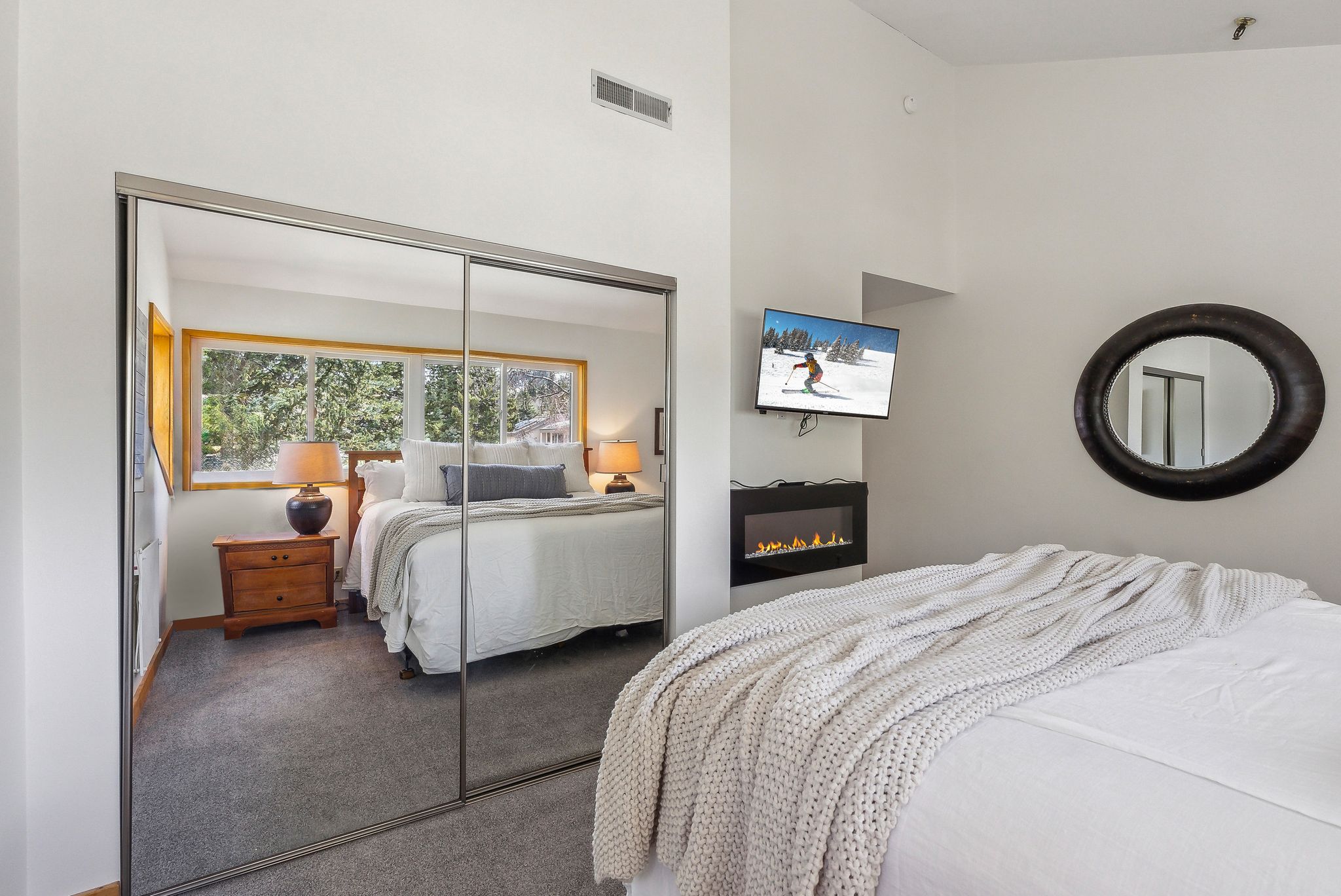 Mirrored closet doors reflecting a bright window, accompanied by an electric fireplace and wall-mounted television