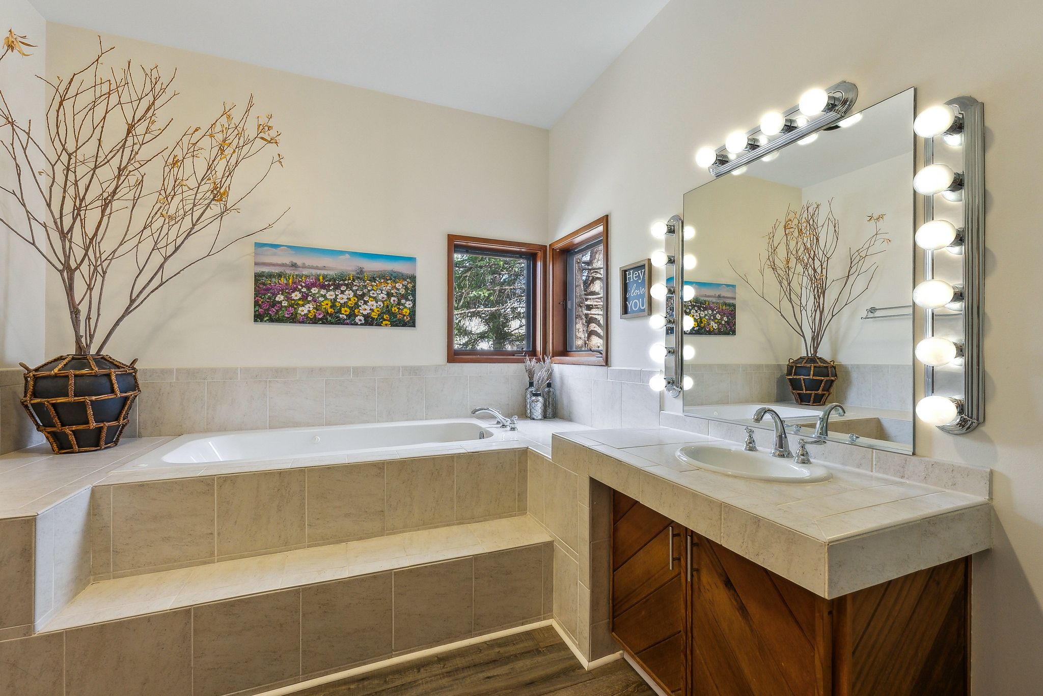 Built-in soaking tub with tiled surround, wood-frame window, vanity with integrated sink, wood-finish cabinetry, and Hollywood-style vanity lighting