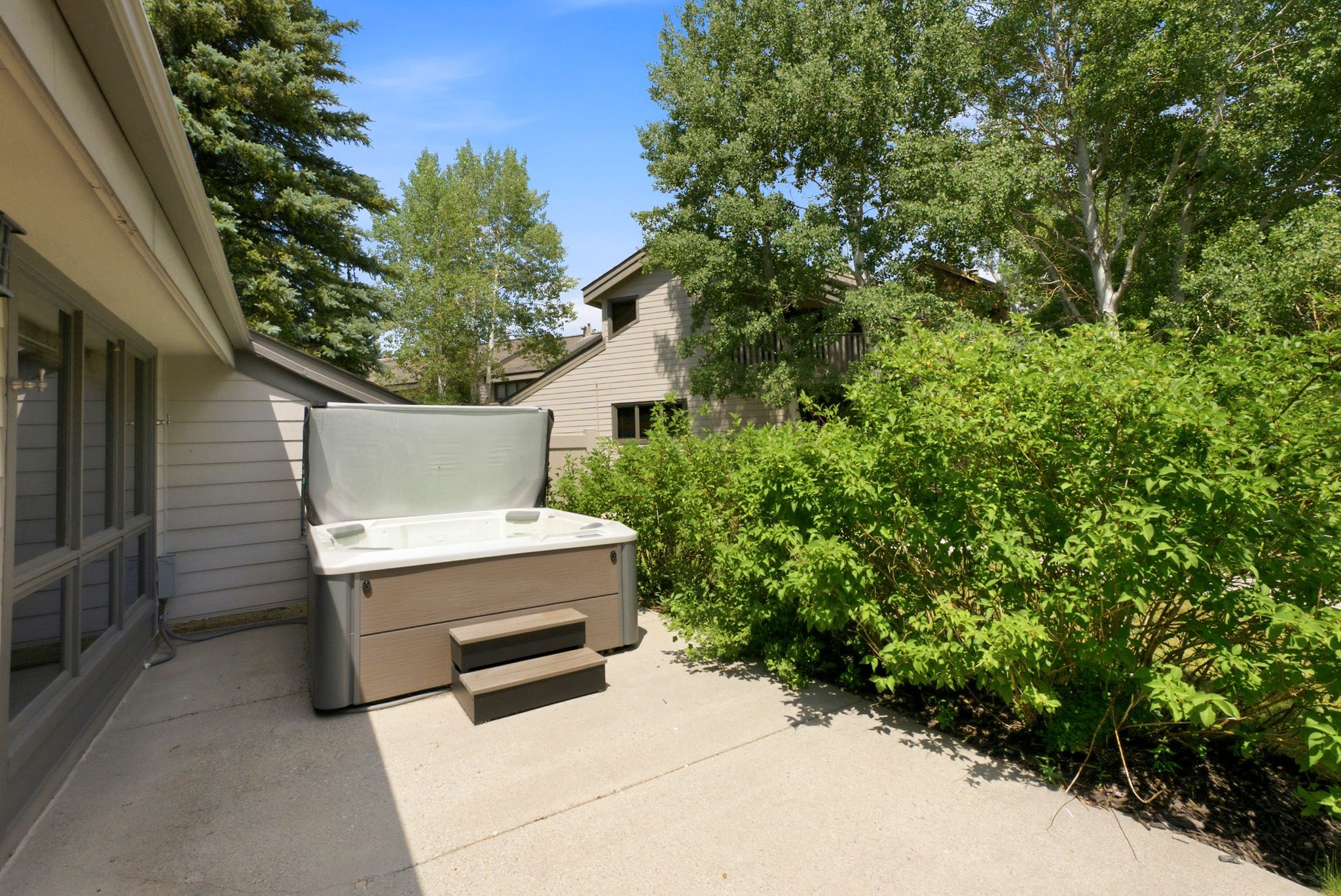 Outdoor patio featuring a hot tub with steps, surrounded by mature trees and lush green shrubs