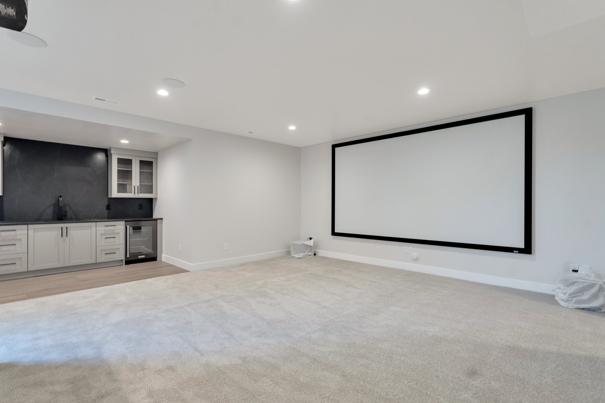 Spacious entertainment room featuring a large projection screen, recessed lighting, and a built-in wet bar with dark countertops, light cabinetry, and a beverage refrigerator