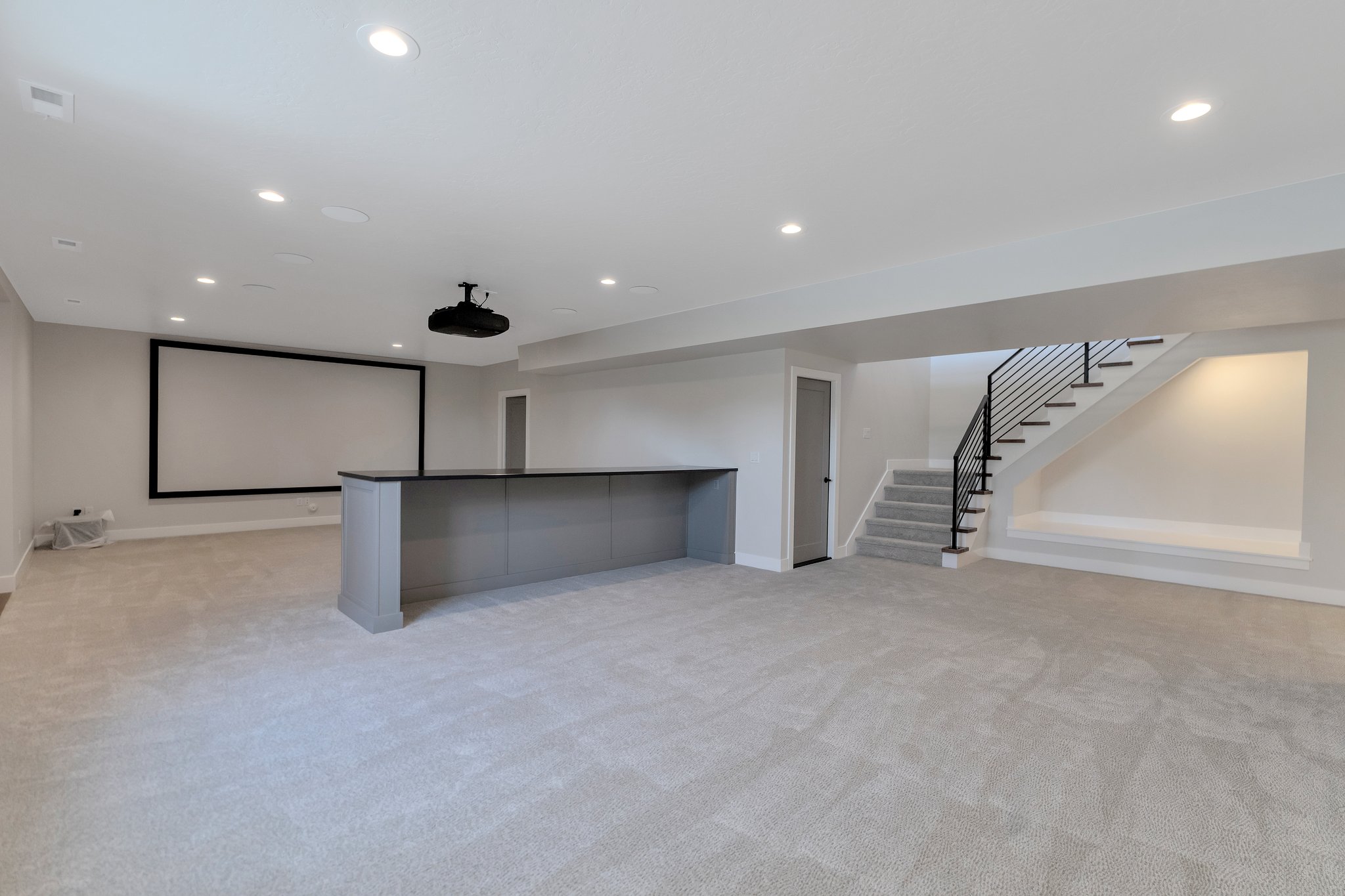 Expansive lower level featuring a built-in projector and large screen, a bar area with dark countertop, light grey carpeting, recessed lighting, and an open staircase with black metal railings