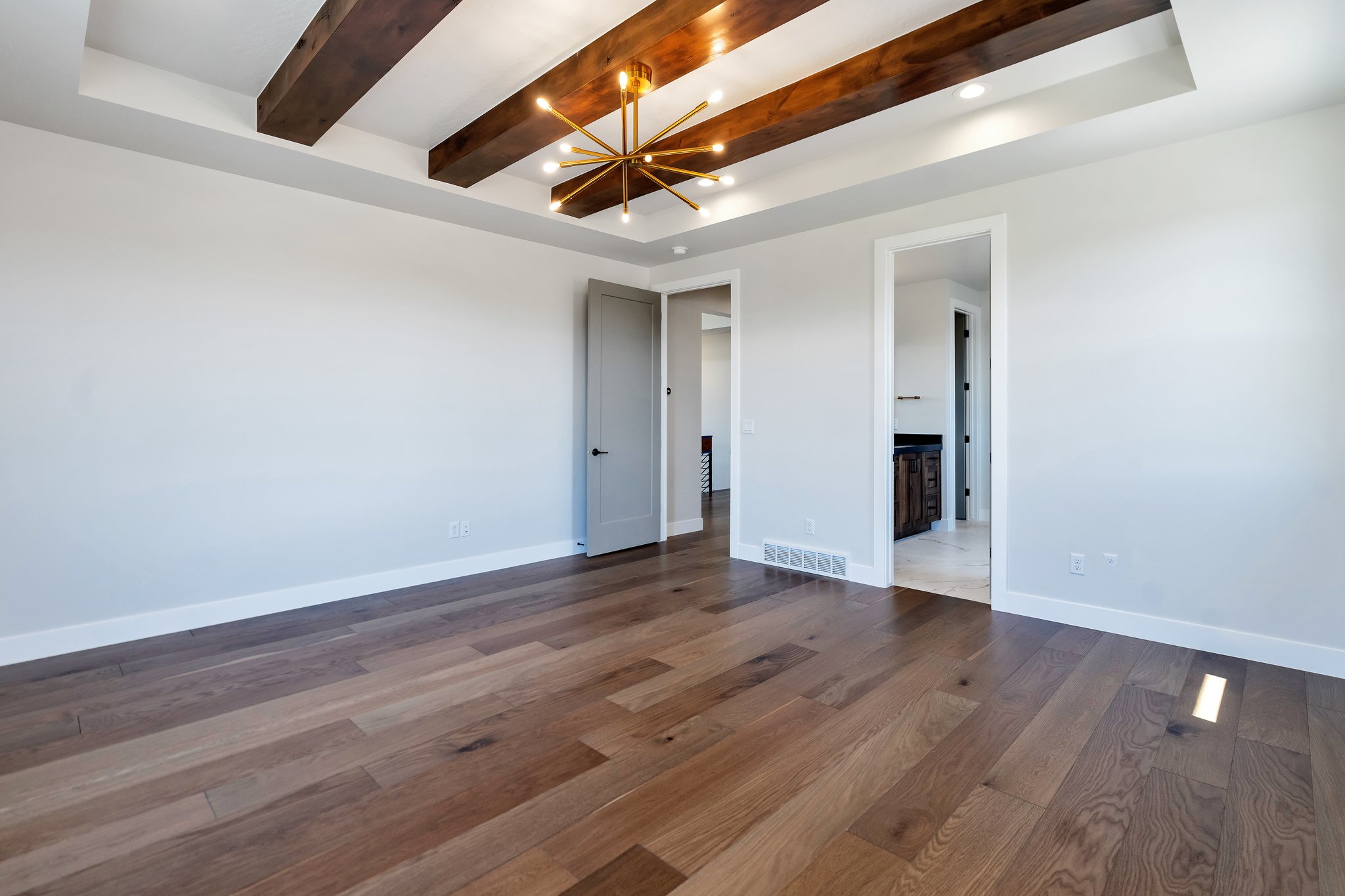 Spacious room featuring wood-finish flooring, a coffered ceiling with dark wood beams, recessed lighting, and a contemporary brass chandelier
