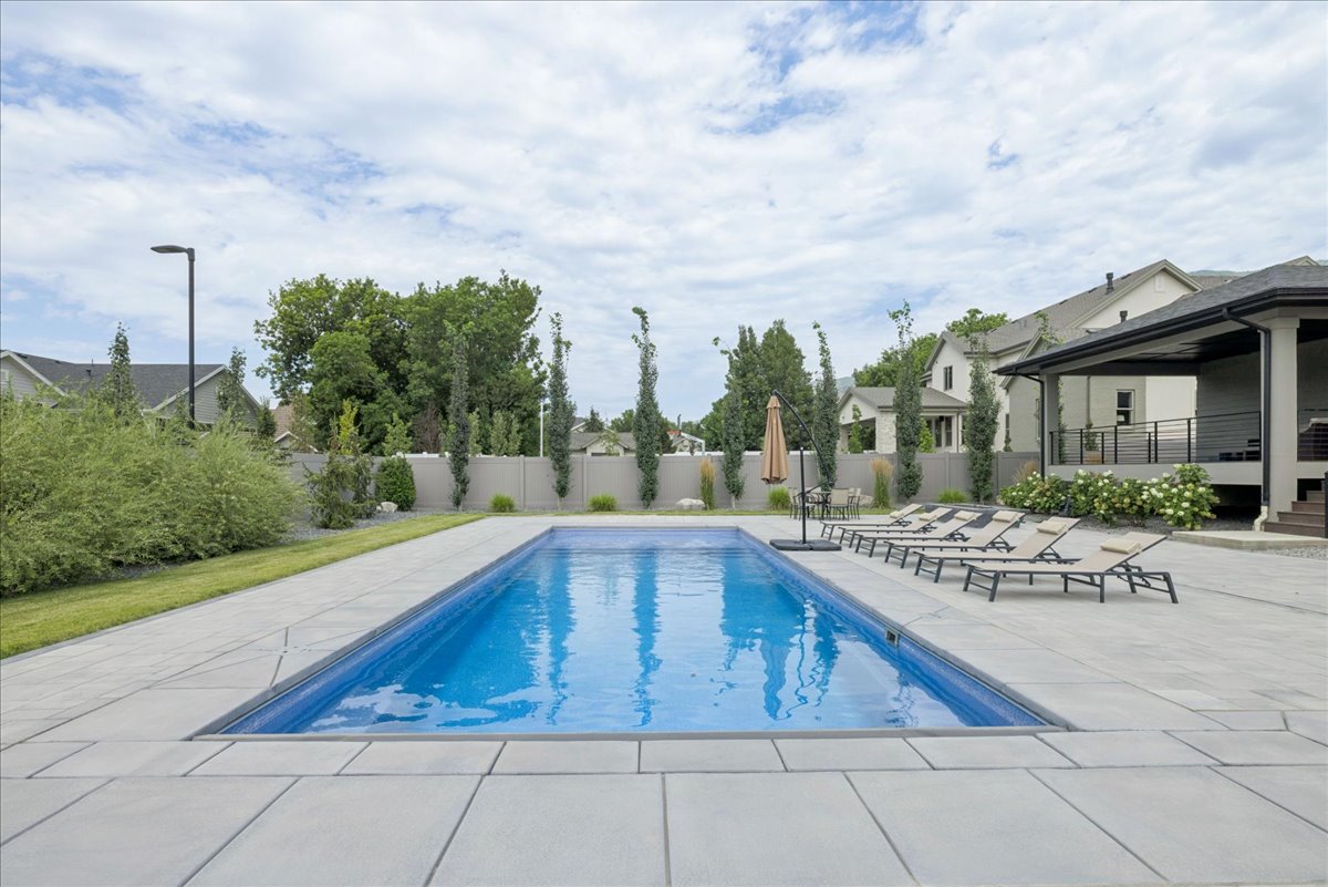 Rectangular swimming pool with expansive light-toned paver surround, bordered by mature trees and a privacy fence