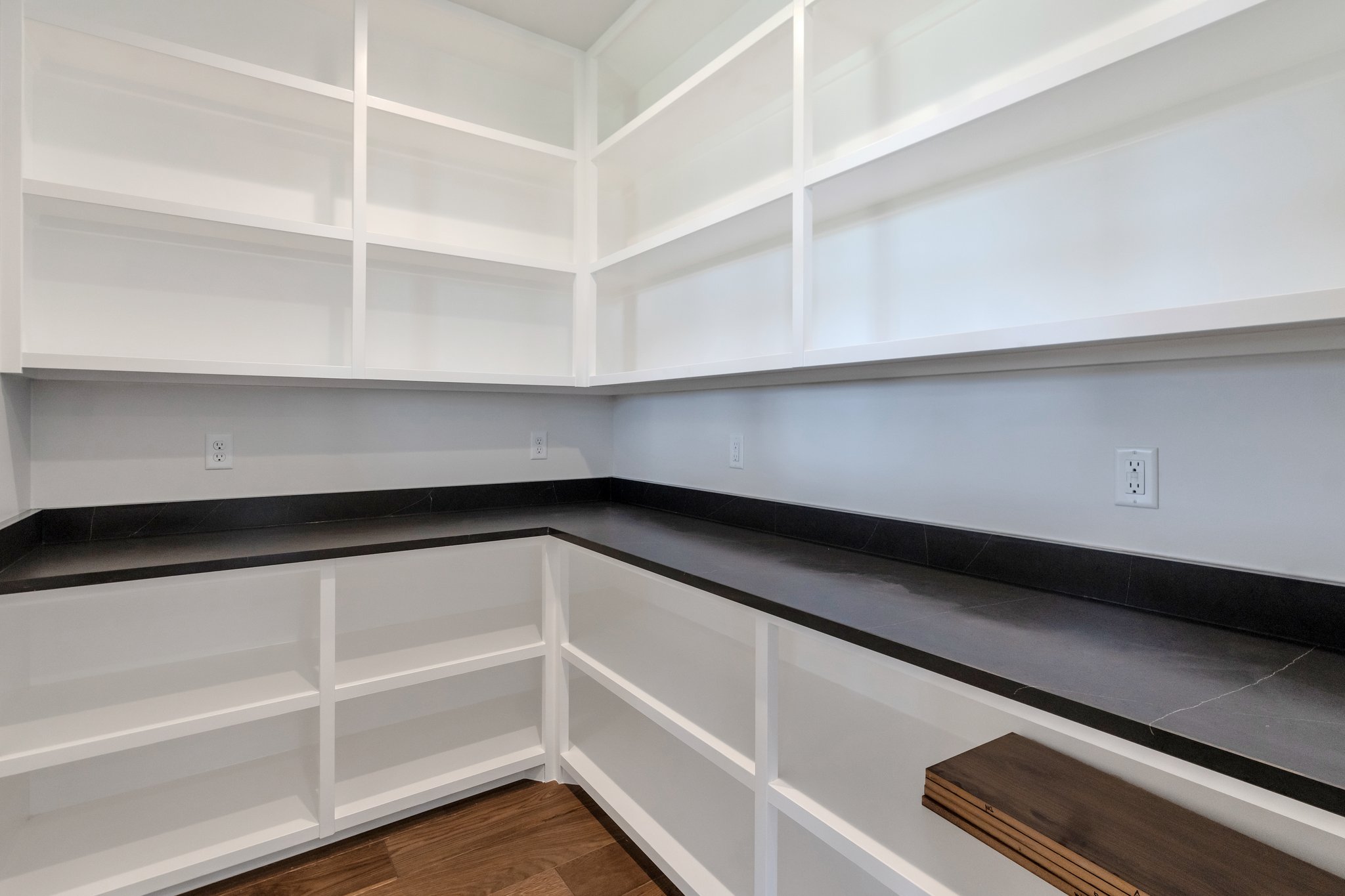 Expansive walk-in pantry featuring white built-in shelving, dark countertops, and wood-finish flooring