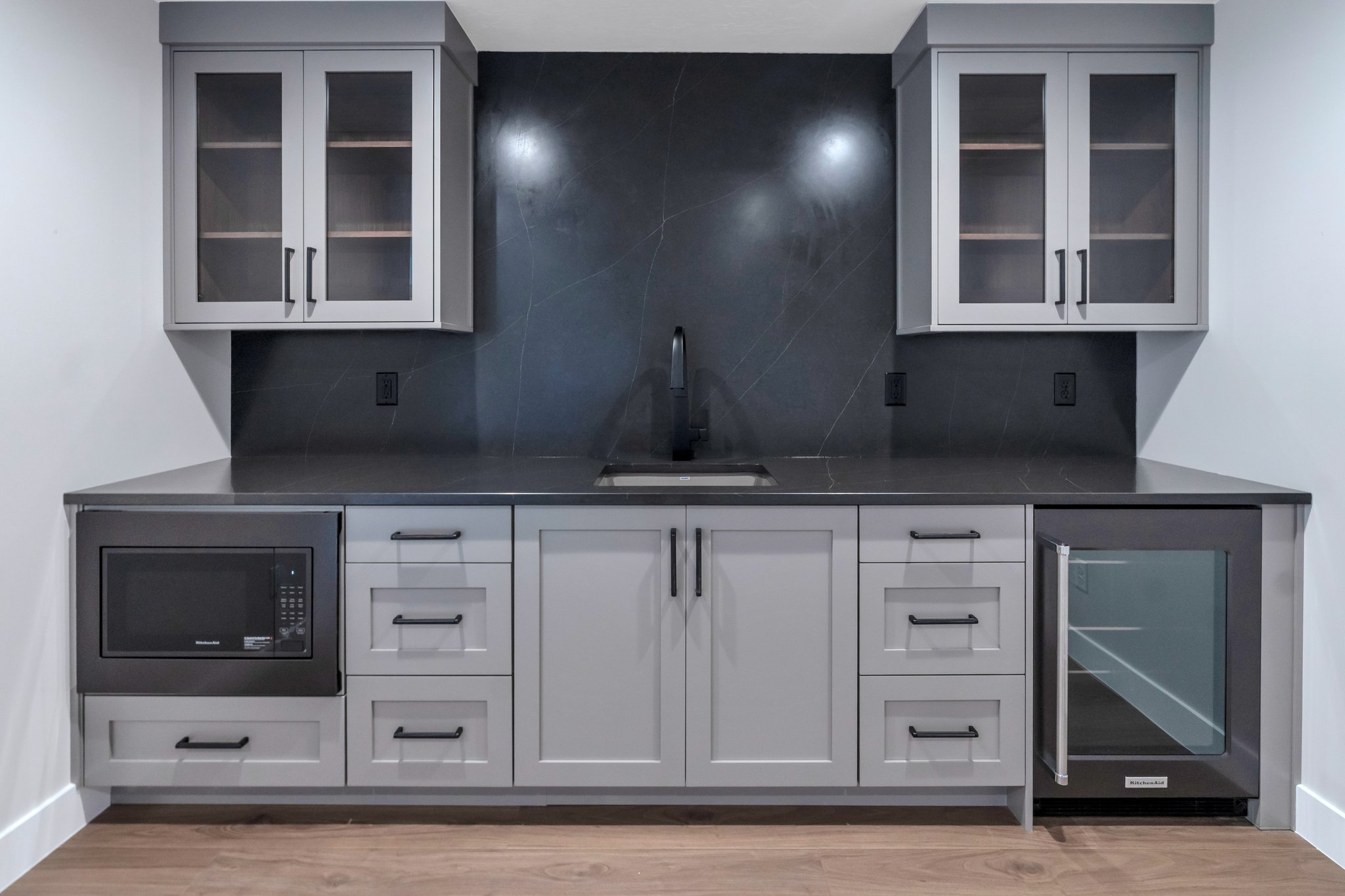 Contemporary wet bar featuring light gray cabinetry with dark hardware, dark countertop, and a dark tile backsplash