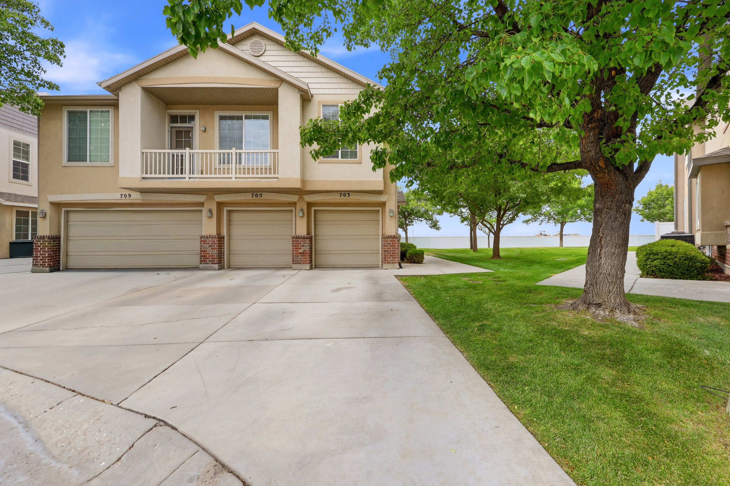 Townhome exterior featuring a stucco finish, a second-story balcony with white railing, individual garages with brick accents, concrete driveways, and a grassy lawn with mature trees