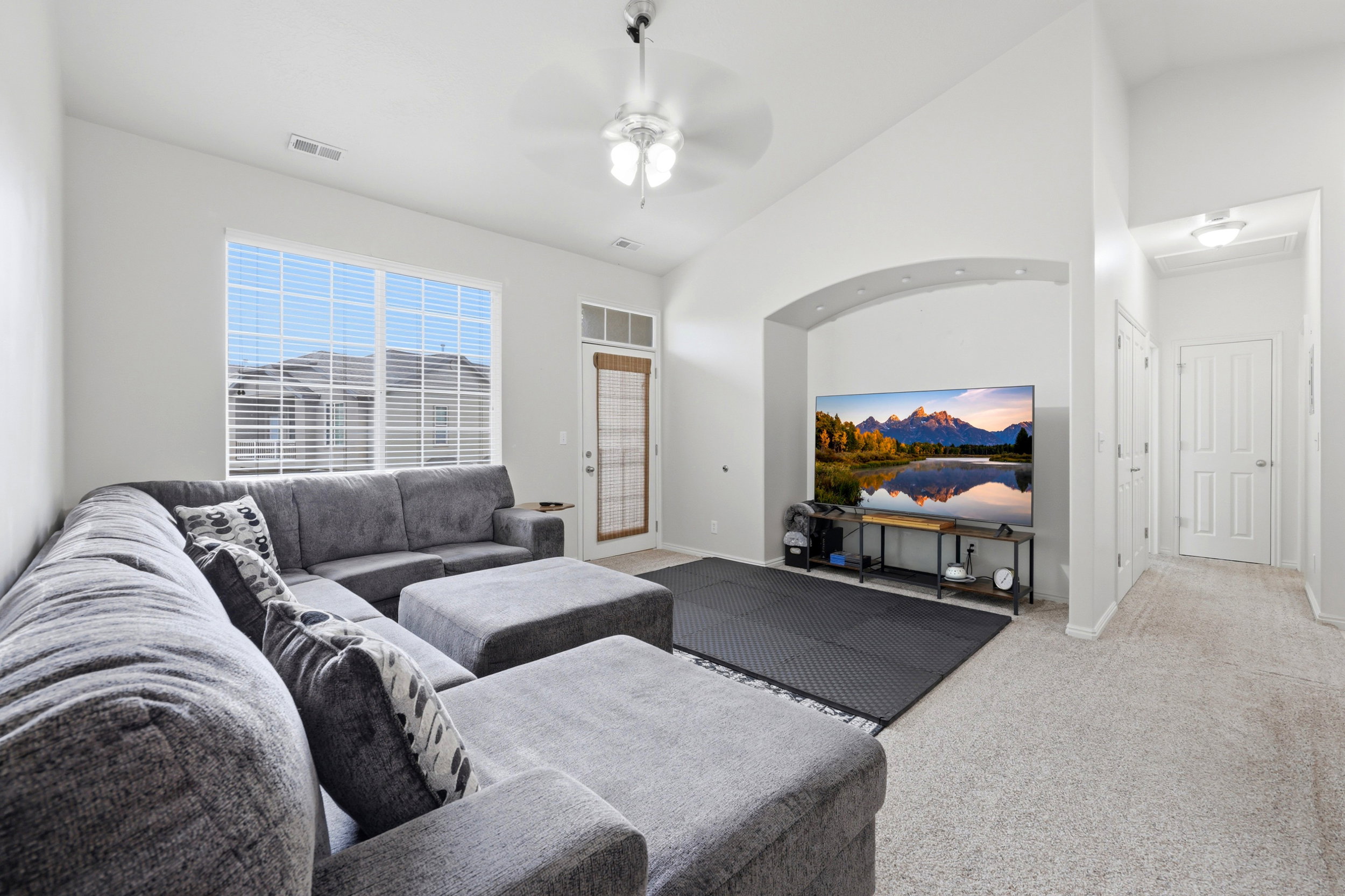 Open-concept living space featuring high vaulted ceilings, large windows with blinds, a ceiling fan, an arched alcove, and light-toned carpeting throughout