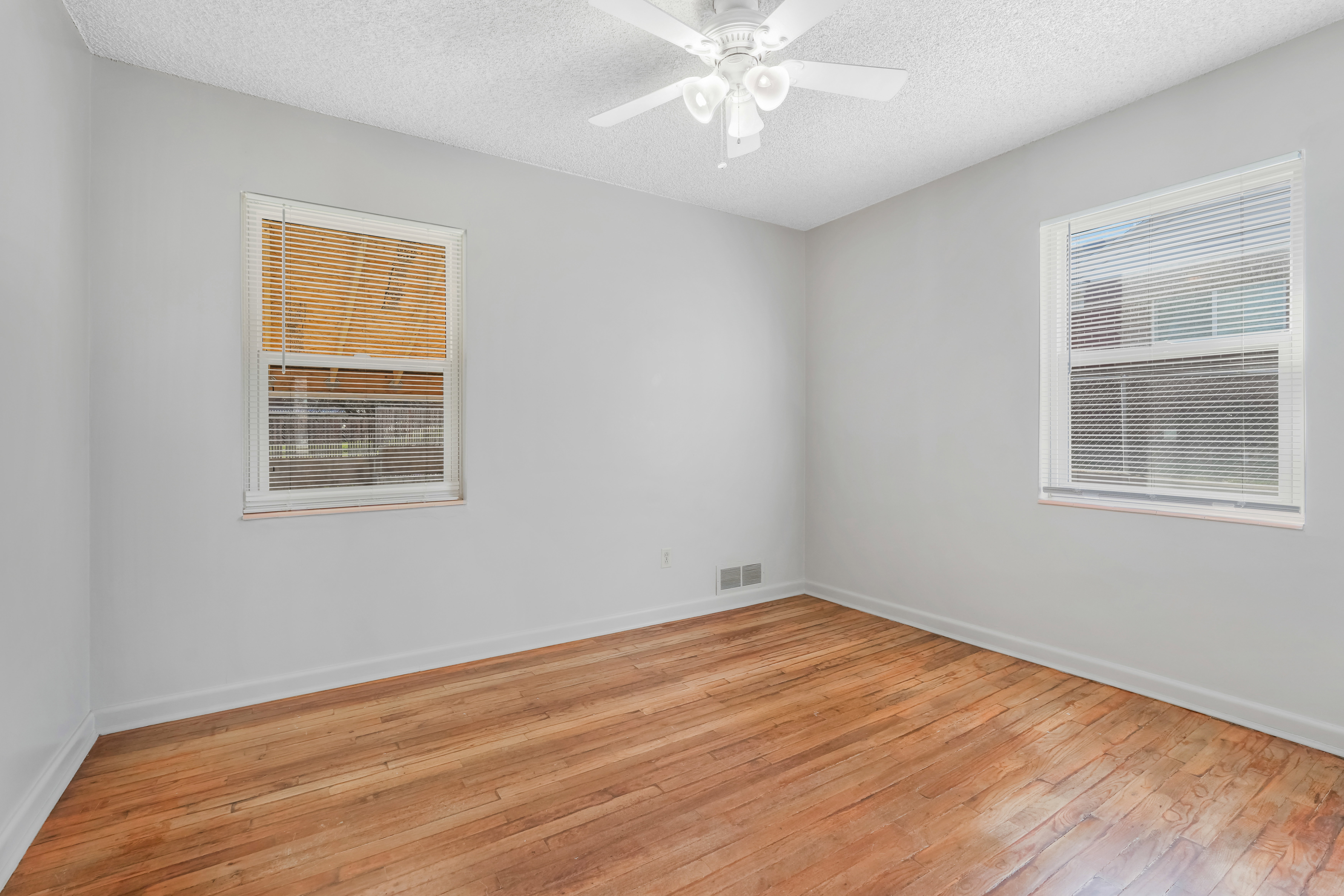 Room with wood-finish flooring, two windows with blinds, a white ceiling fan, and light gray painted walls