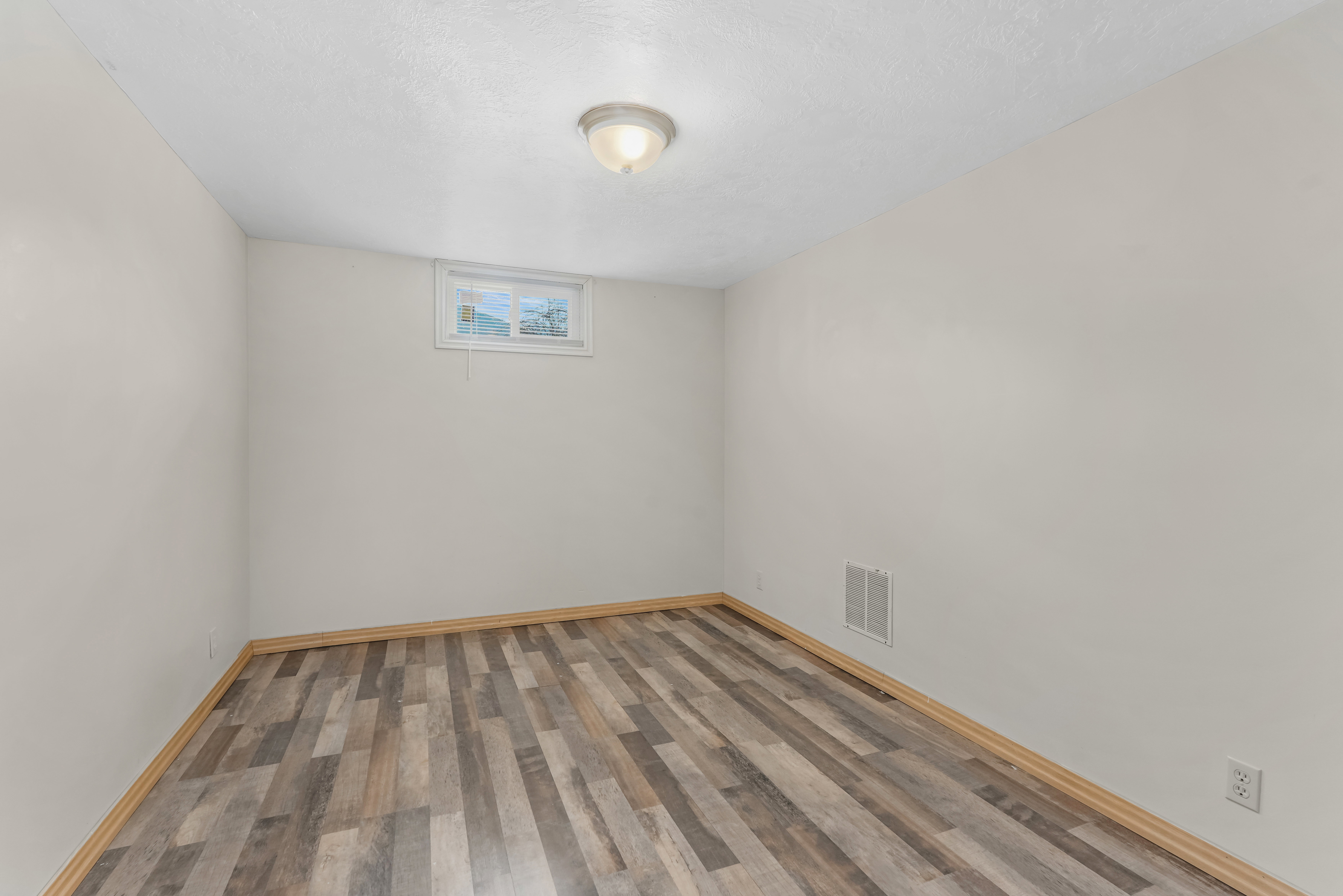 Neutral-toned room with multi-hued wood-finish flooring, light baseboards, and a single window providing natural illumination