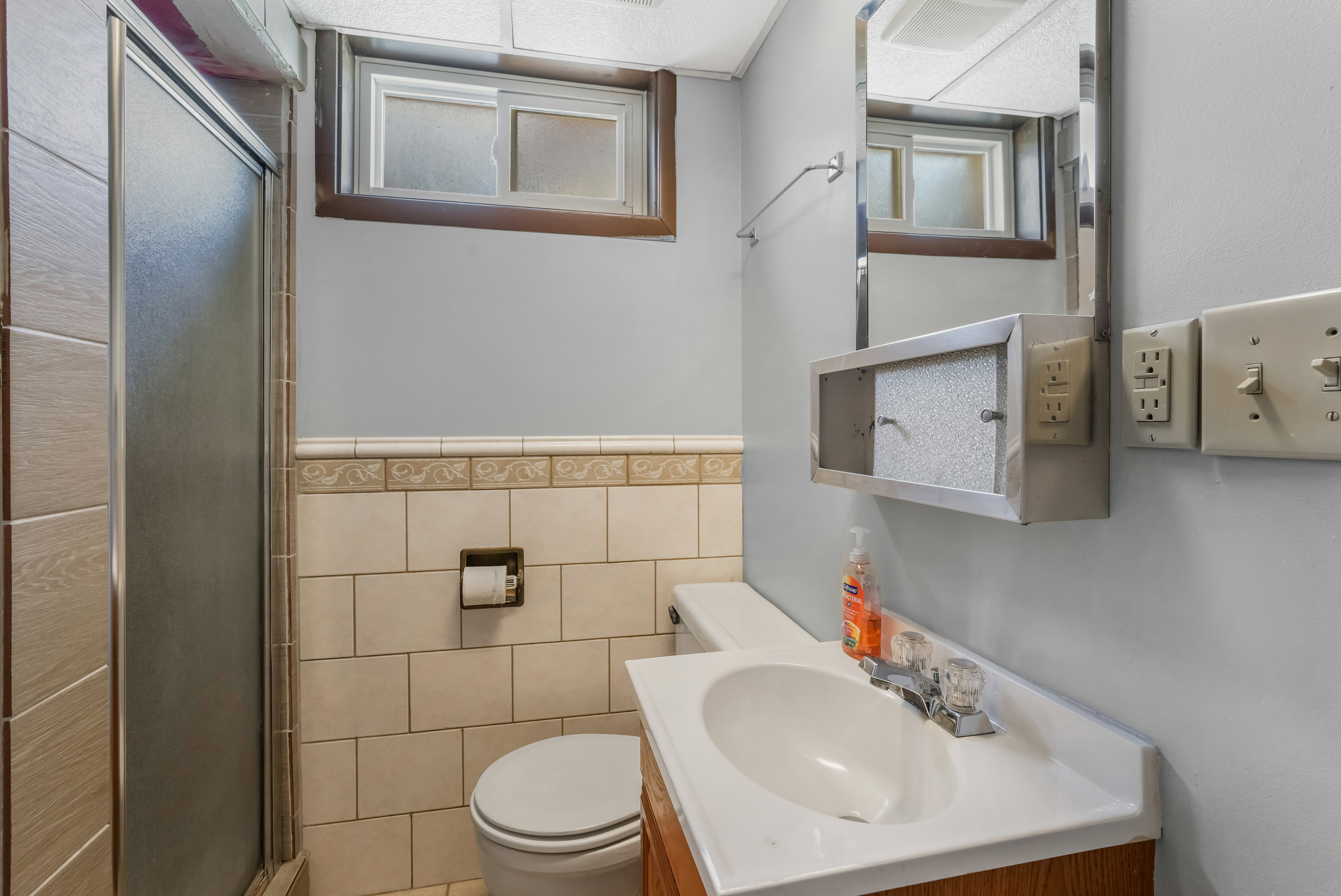 Bathroom featuring a glass-enclosed shower, wall-mounted vanity with integrated sink, toilet, tile wainscoting, and a mirrored medicine cabinet