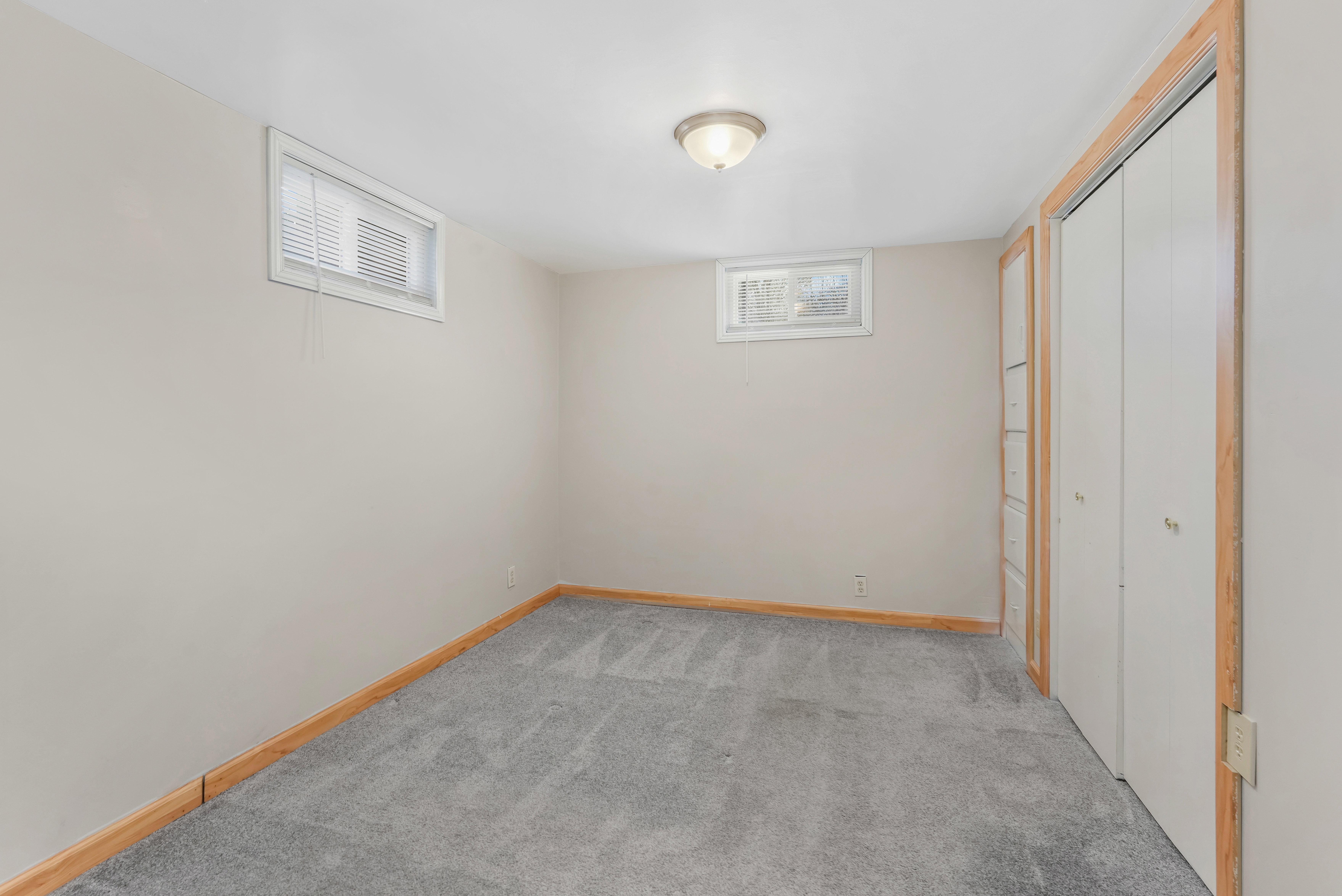 Room featuring neutral wall paint, gray carpet flooring, wood-finish trim, a flush-mount ceiling light fixture, and two windows with blinds