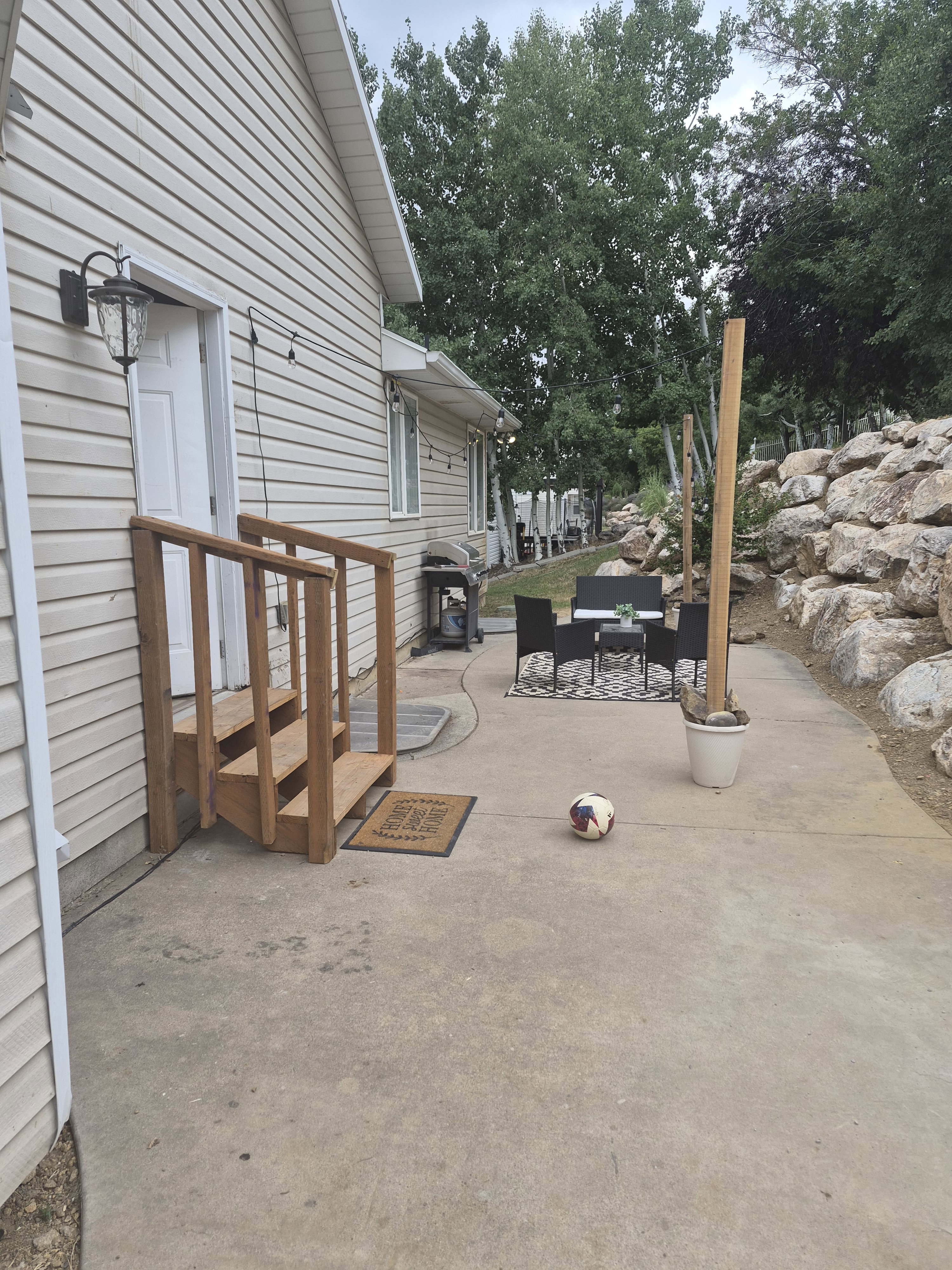 Concrete patio featuring a wooden staircase with railing, exterior door with mounted light fixture, and a built-in rock wall