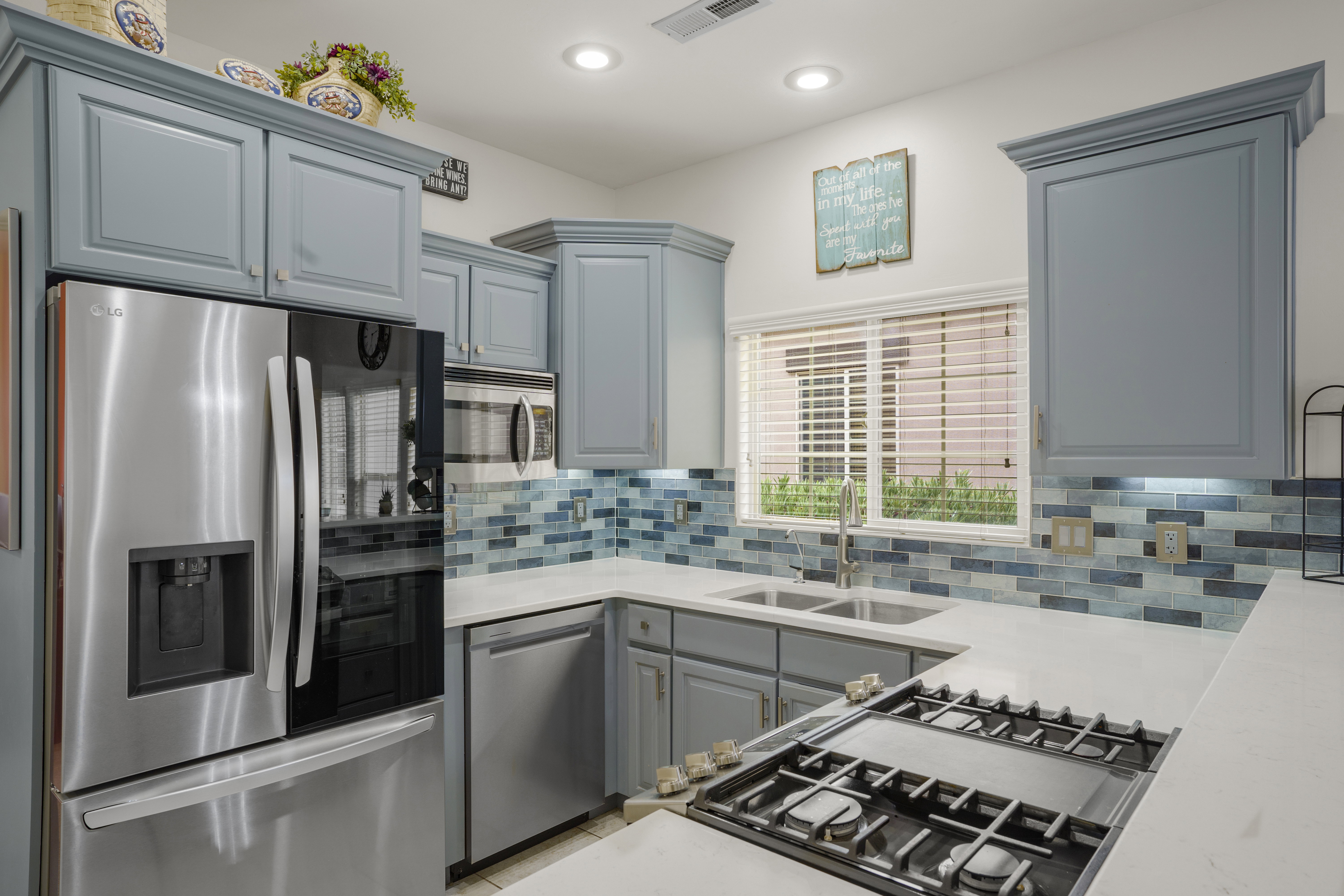 Kitchen featuring light blue cabinetry, stainless steel appliances, a tile backsplash with blue and white tones, white countertops, and a window with blinds