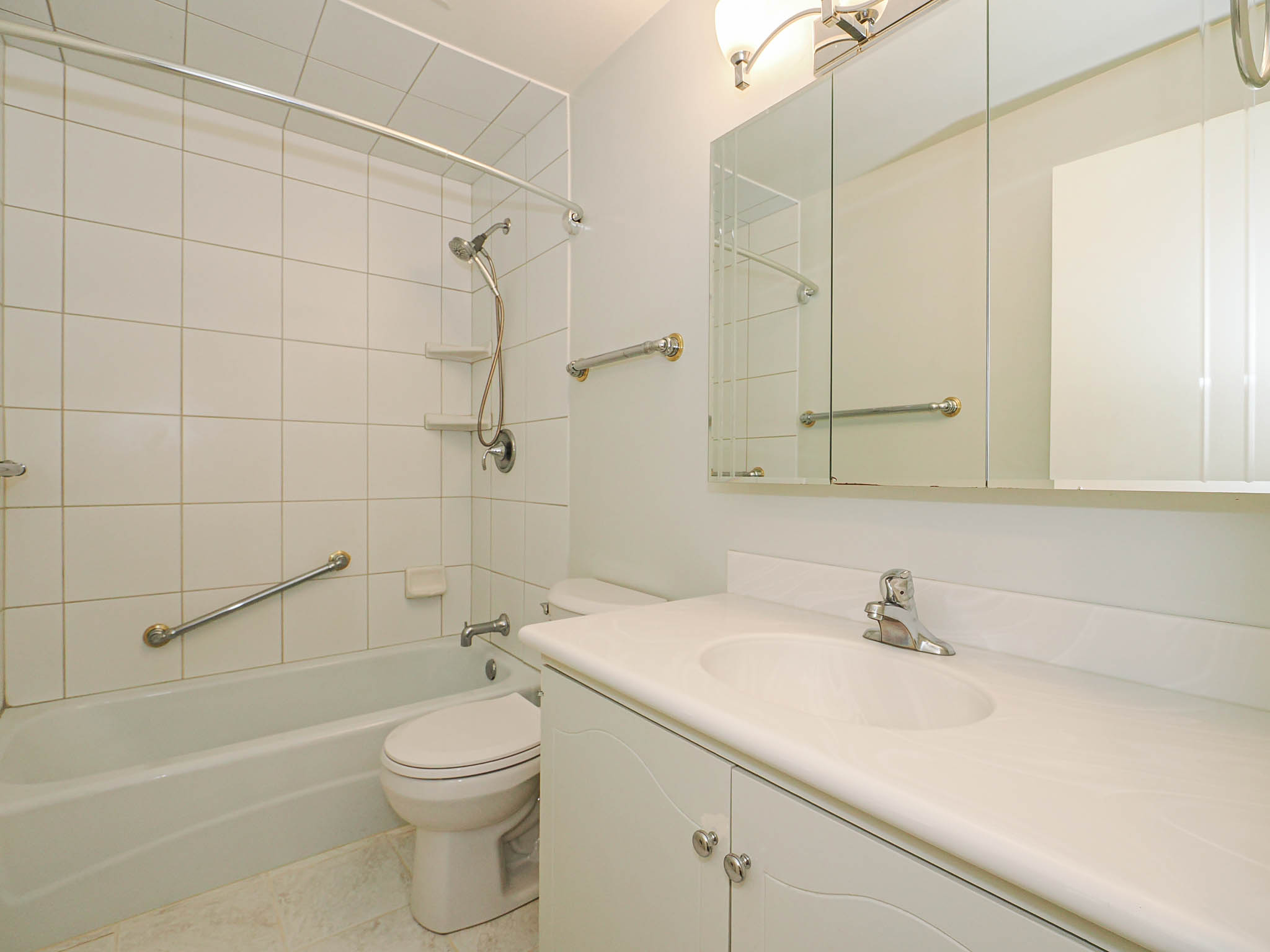 Bathroom featuring a white bathtub with subway tile surround, white vanity with integrated sink, tri-fold mirrored medicine cabinet, and overhead light fixture