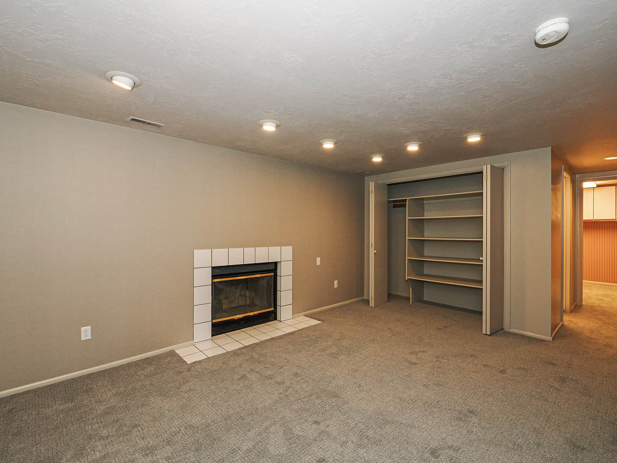 Recessed lighting illuminating a plush carpeted room with a fireplace featuring a white tile surround and hearth