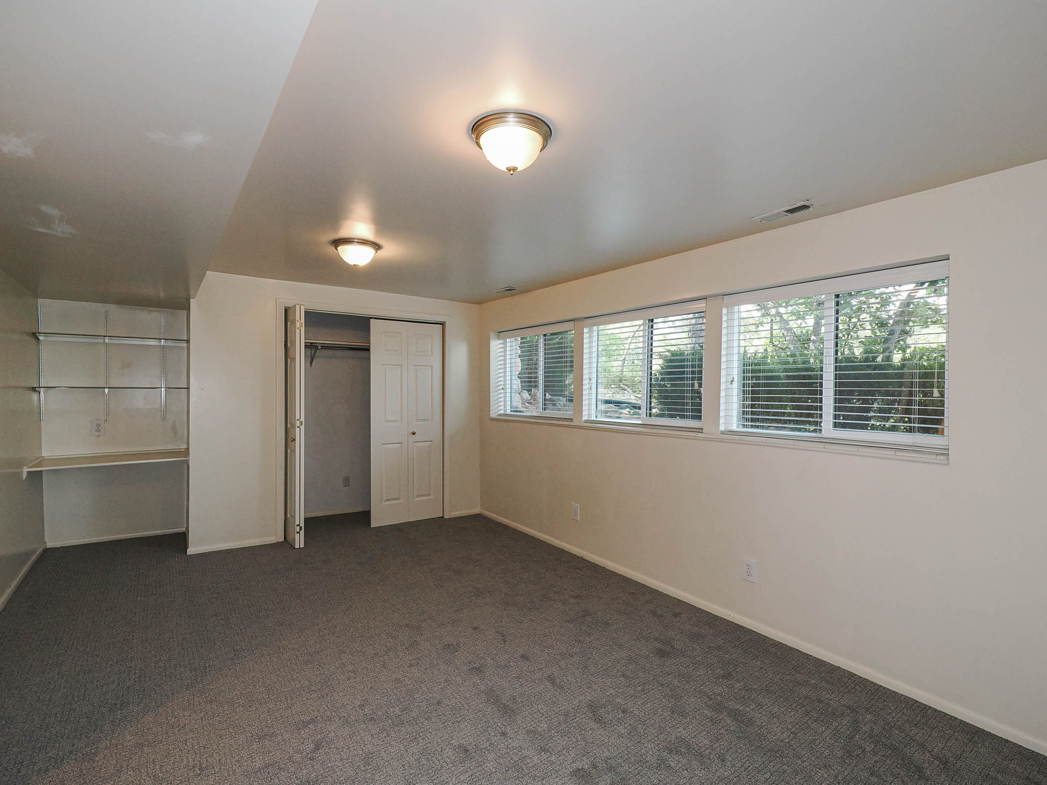 Spacious room featuring carpeting, multiple windows with blinds, a closet with bi-fold doors, built-in shelving, and two ceiling-mounted light fixtures
