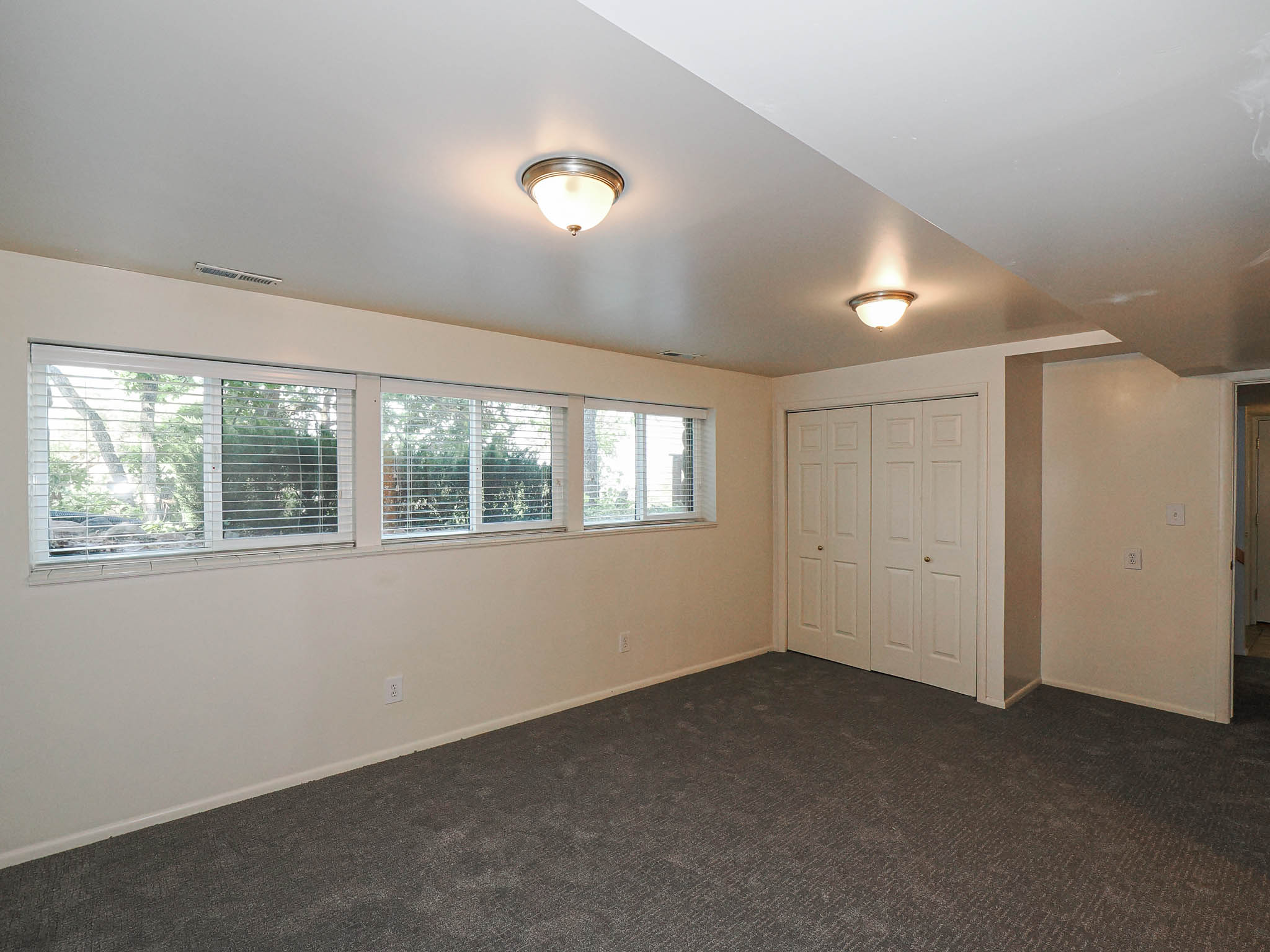 Spacious room featuring new gray carpeting, multiple horizontal windows with blinds, a double bifold closet, two ceiling-mounted dome light fixtures, and neutral wall paint