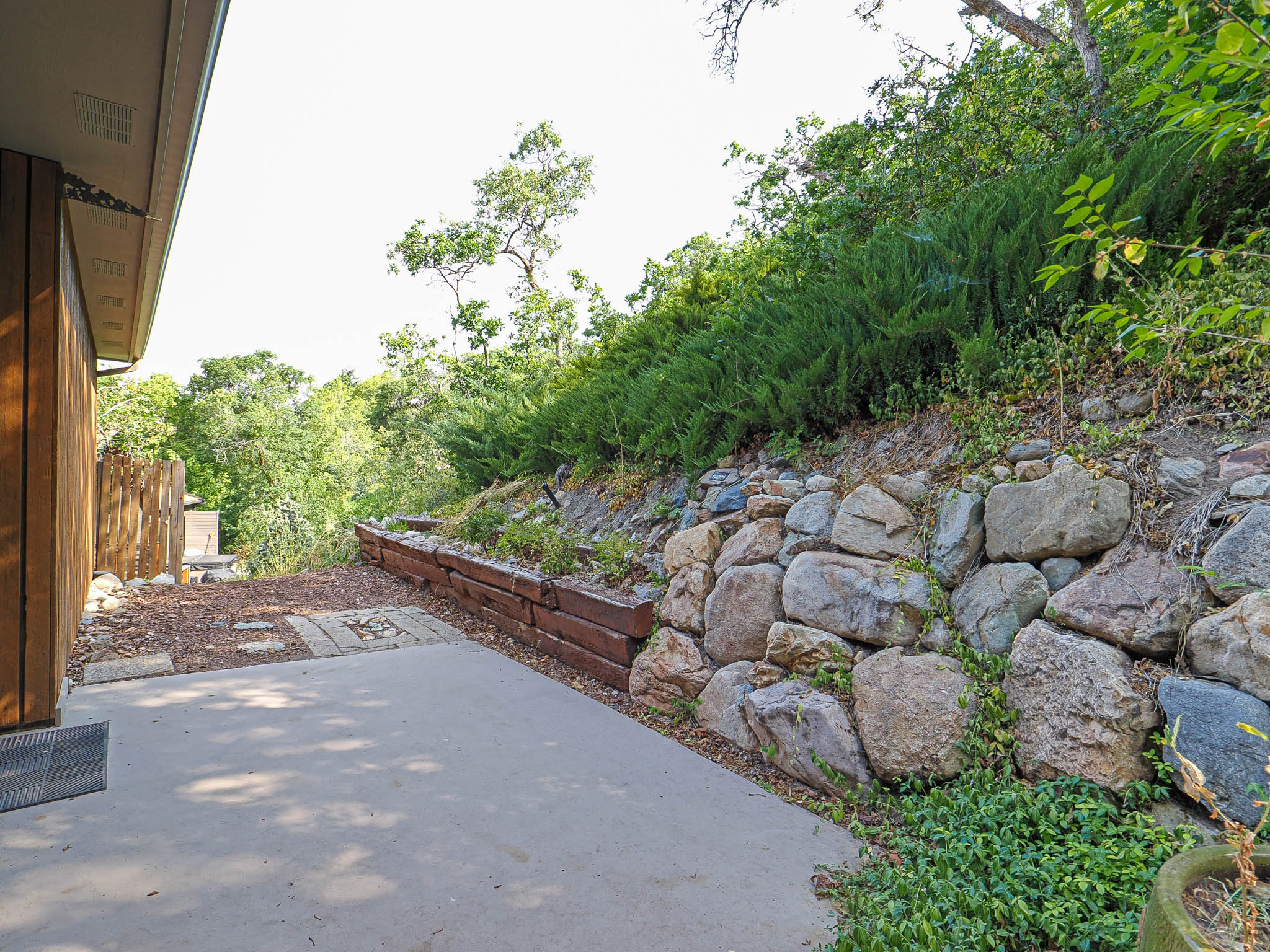 Outdoor patio with concrete surfacing, bordered by a stone retaining wall and an upper tier of natural landscaping