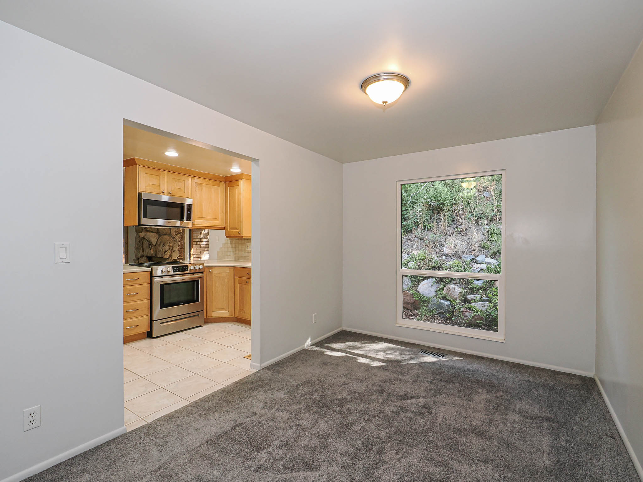 Carpeted room with a single window, featuring a recessed kitchen area with wood cabinetry, stainless steel appliances, and tile flooring