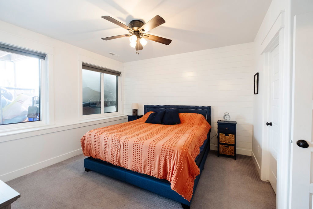 Bright bedroom featuring two windows with roller shades, white shiplap accent wall, grey carpeting, and a ceiling fan with integrated lighting