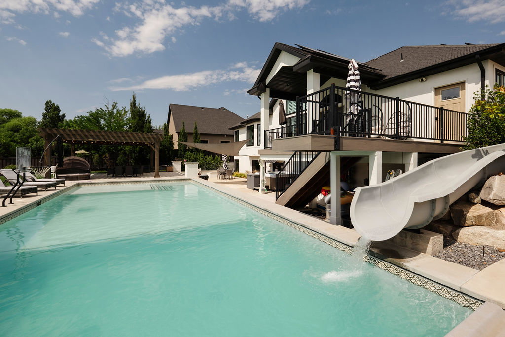 Rectangular swimming pool featuring a tiled waterline, a built-in slide, a pergola structure, a raised patio deck with black railings, and a two-story exterior