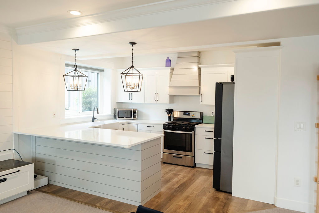 Modern kitchen featuring white shaker cabinetry, stainless steel appliances, a custom range hood, wood-finish flooring, and two geometric pendant light fixtures