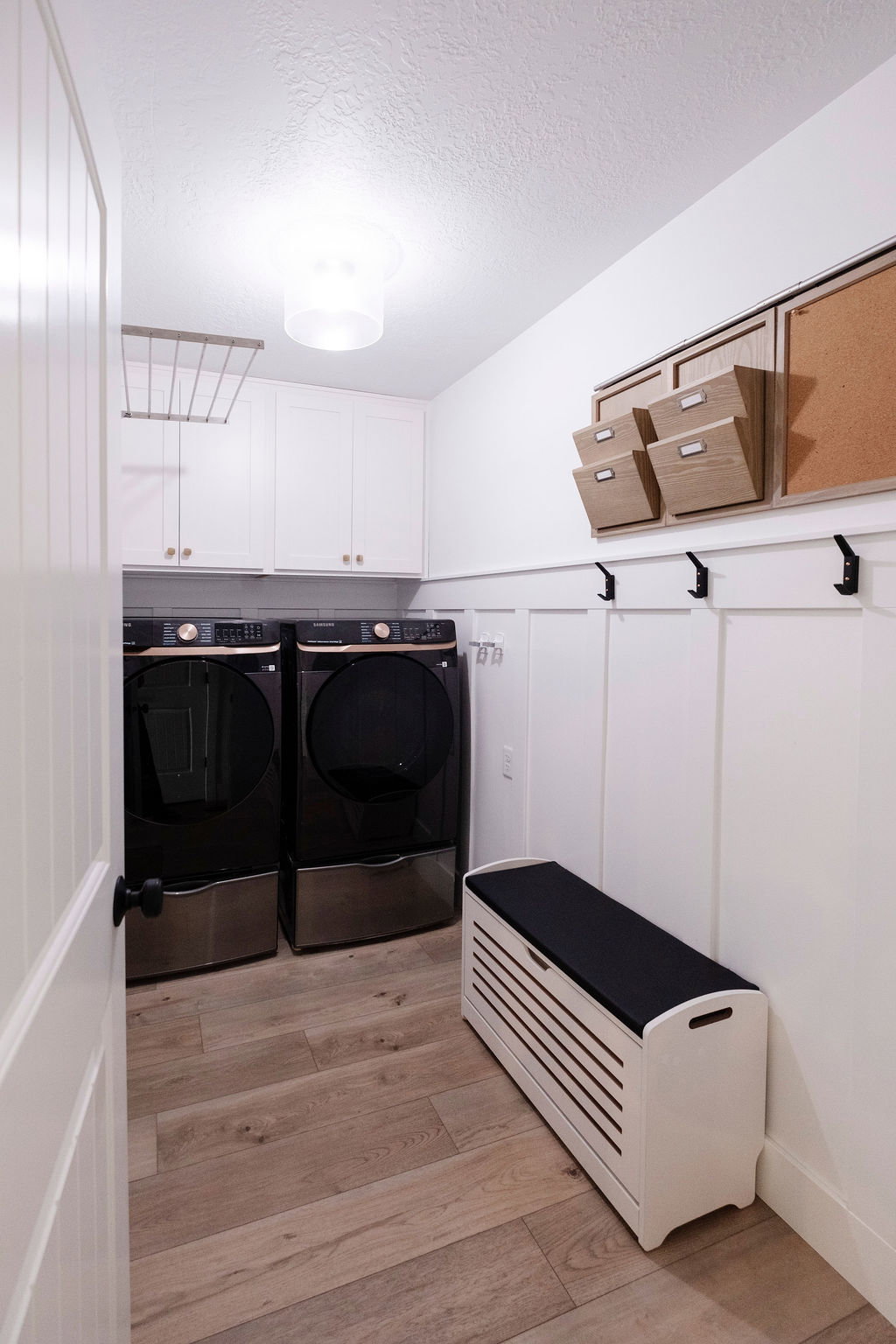 Dedicated laundry room featuring wood-finish flooring, white shaker-style cabinetry, and a board and batten accent wall with black coat hooks