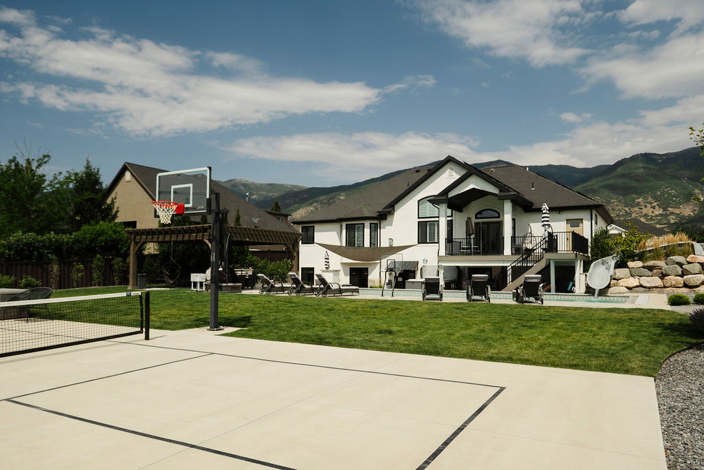 Expansive backyard featuring a multi-sport court with a basketball hoop and net, a manicured lawn, and a large patio area with a swimming pool