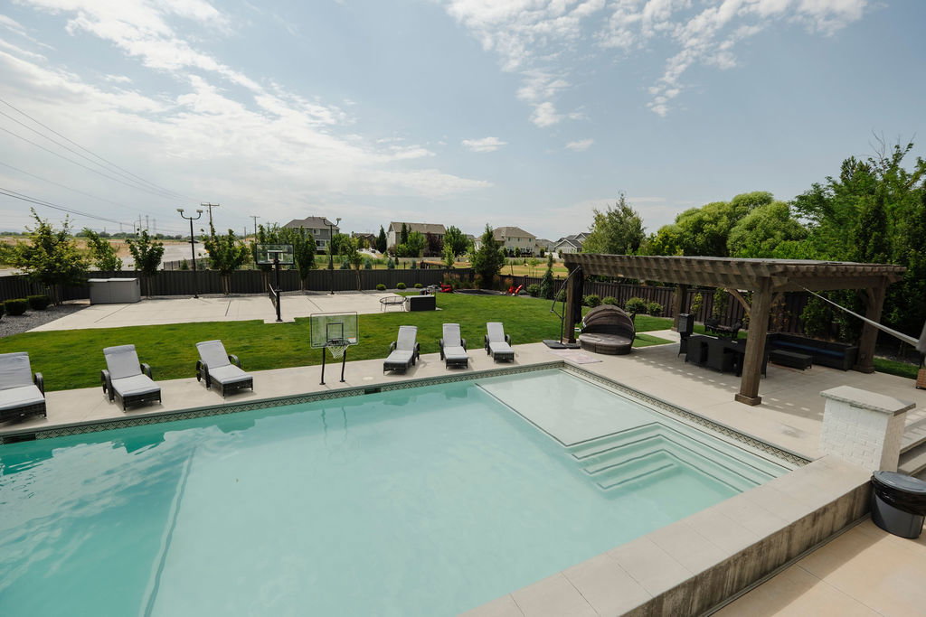 Expansive outdoor living area featuring a rectangular swimming pool with integrated steps, a large paved patio, and a substantial wooden pergola