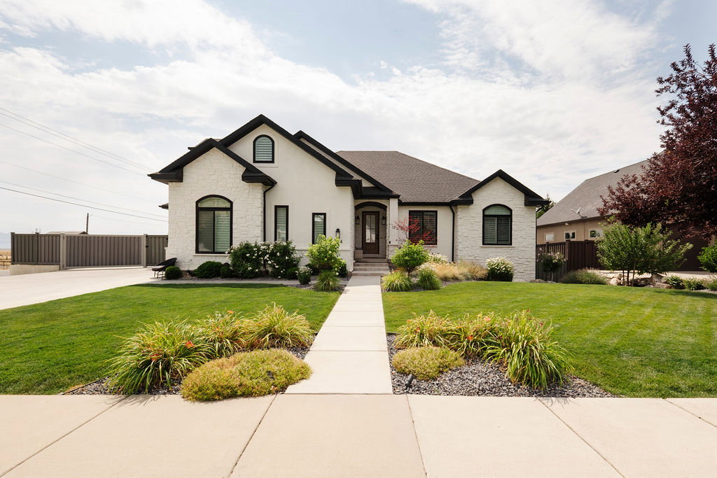 Modern stucco exterior with black trim windows, a dark shingled roof, a concrete walkway, and a manicured lawn with varied landscaping