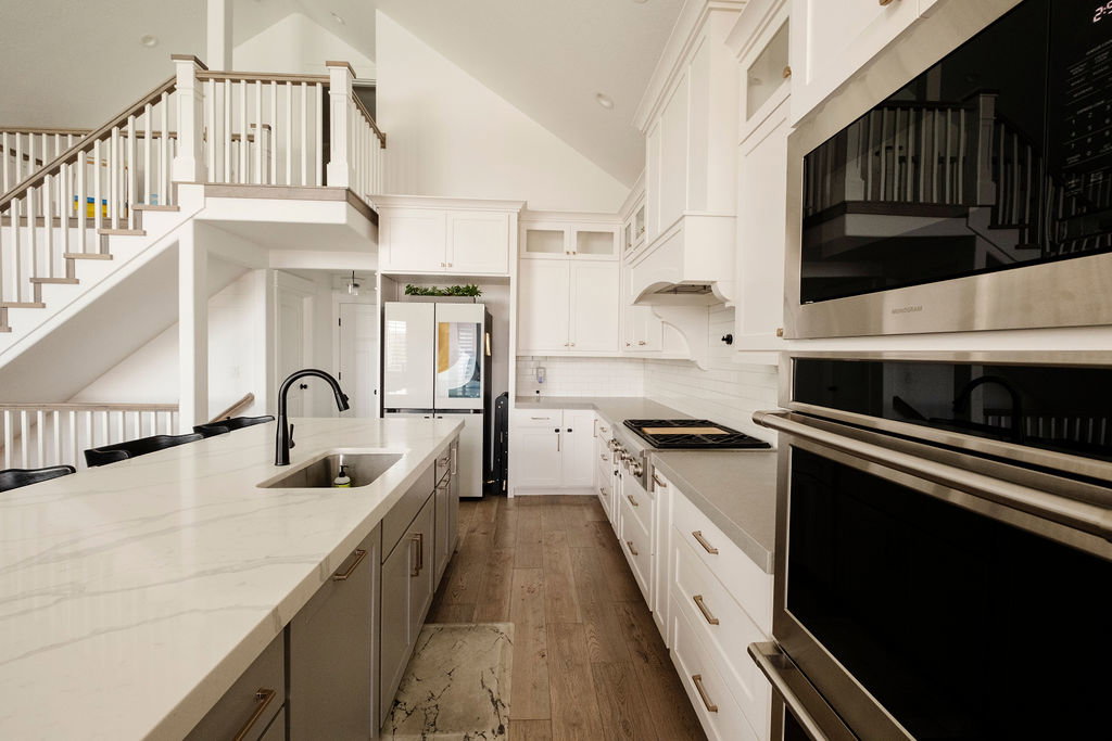 Open-concept kitchen featuring a large island with an undermount sink and matte black faucet, light wood-finish flooring, white upper and gray lower cabinetry, and stainless steel built-in appliances