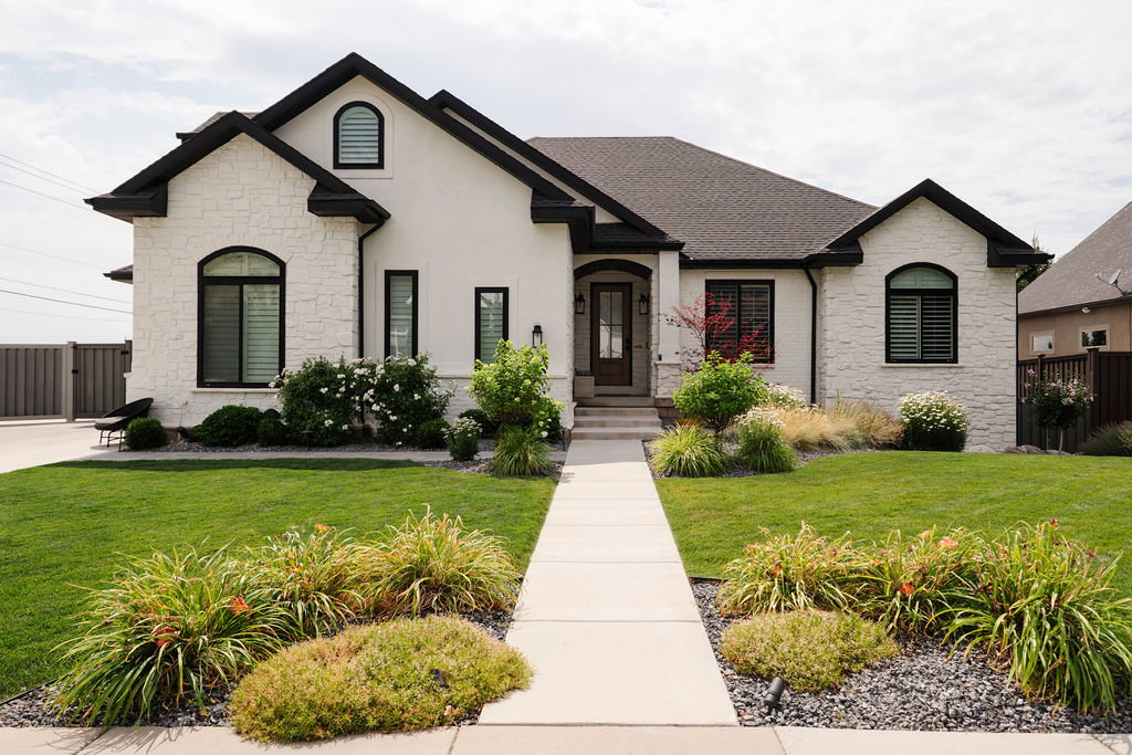 Contemporary residence featuring a blend of white stucco and stone veneer, dark trim accents, a concrete walkway, and a meticulously landscaped front yard with varied plantings