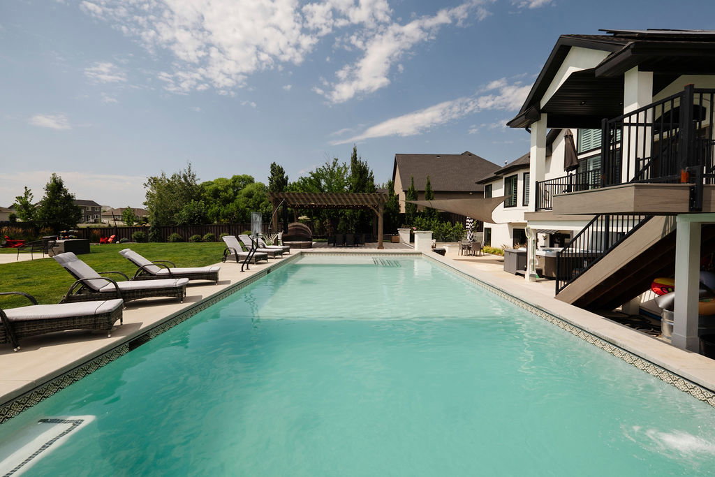 Expansive backyard featuring a rectangular swimming pool with integrated steps, a surrounding light-toned paved patio, and a separate elevated deck with exterior stairs