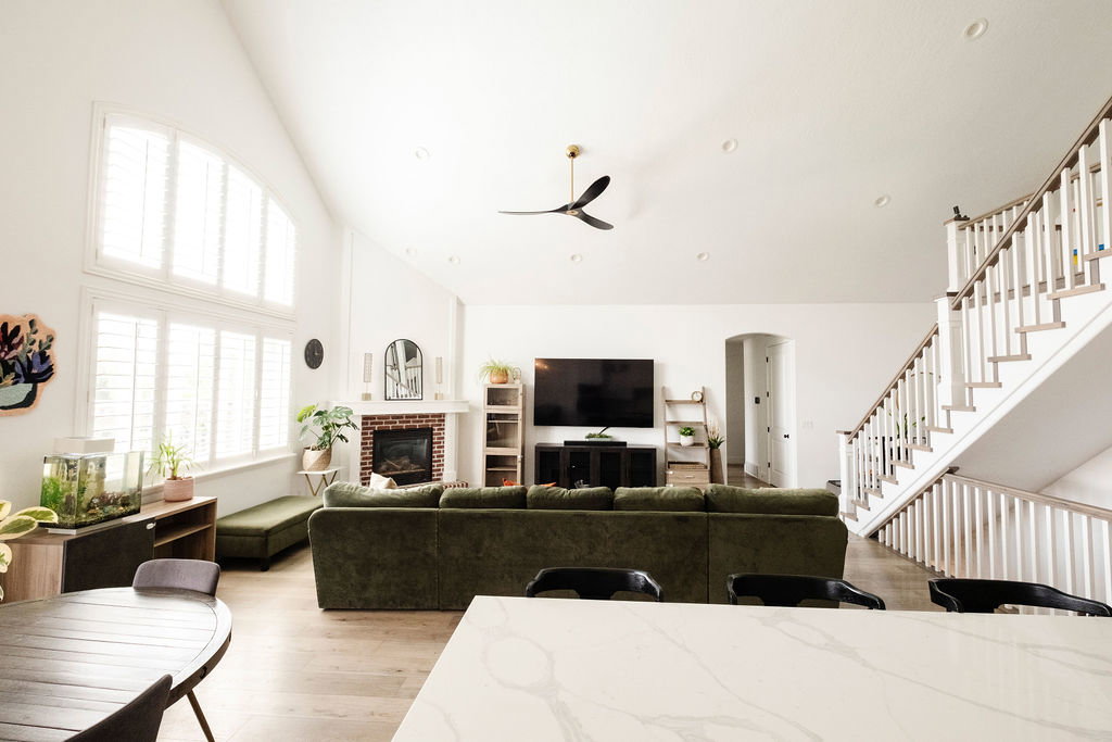 Spacious living area featuring vaulted ceilings, large windows with plantation shutters, a brick fireplace with mantel, recessed lighting, and wood-finish flooring