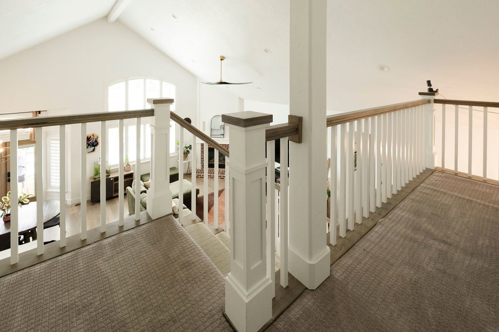 Carpeted upper-level landing with white balustrade and wood-finish handrail overlooking a vaulted great room