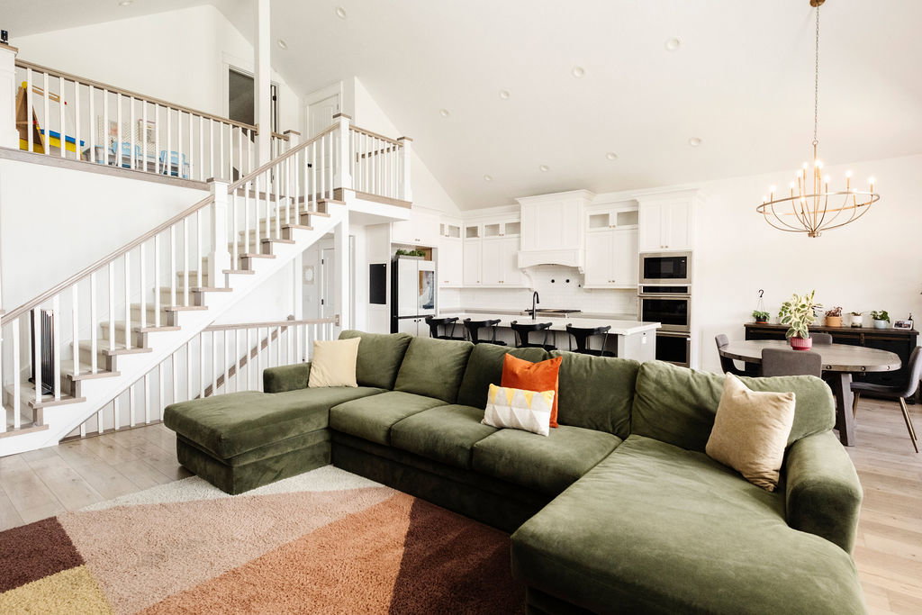 Open concept living area with vaulted ceilings, featuring wood-finish flooring, a two-story staircase with white balusters, a kitchen with white cabinetry and built-in double ovens, and a dining area with a contemporary chandelier