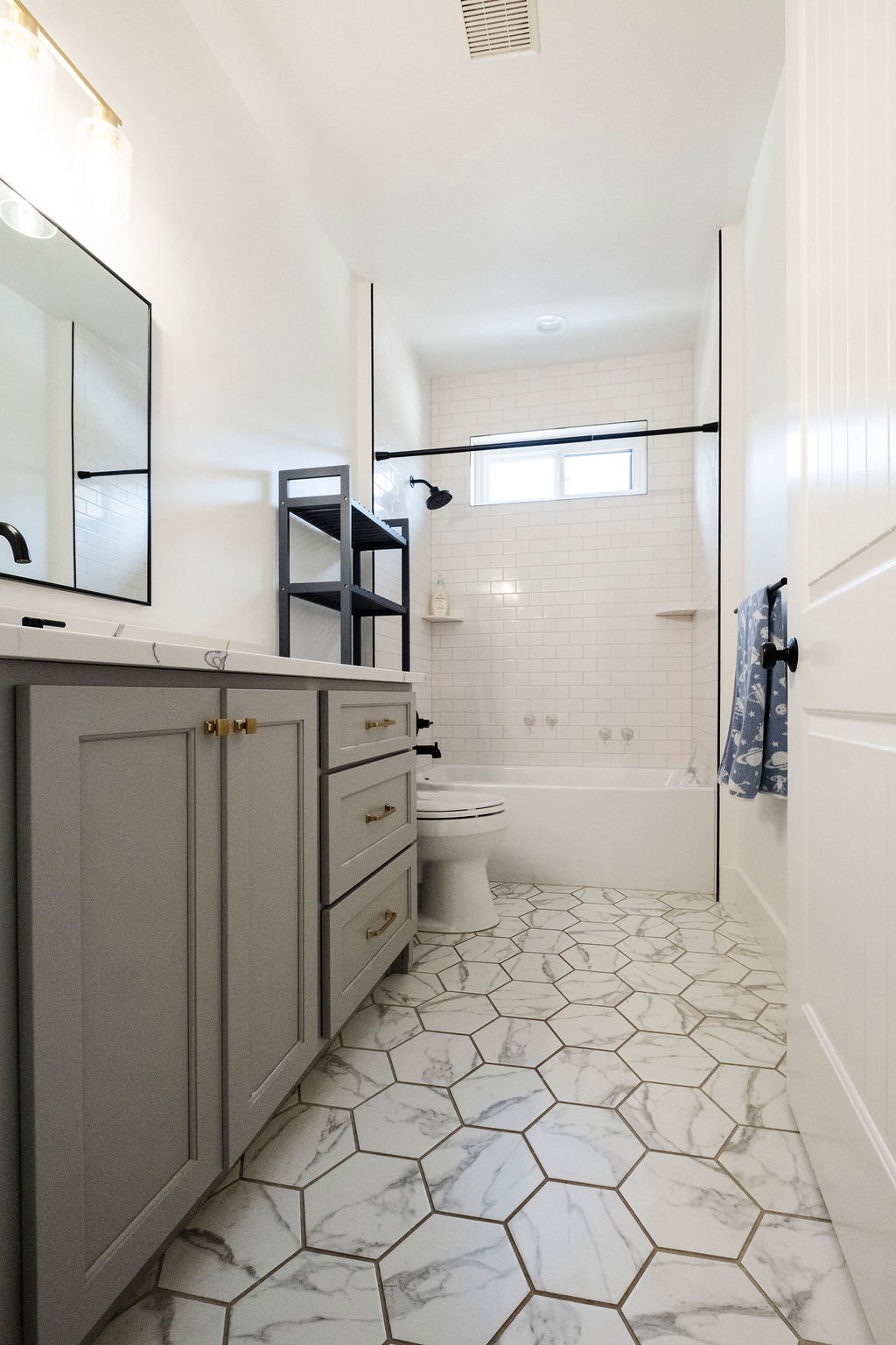 Bathroom featuring hexagon marble-look floor tiles, a light gray vanity with gold-tone hardware, a white subway tile bath surround, and a tub with a black shower fixture