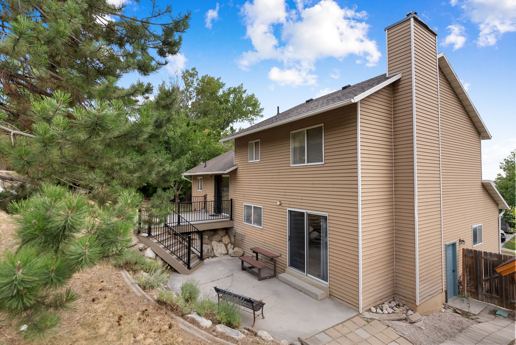Rear exterior featuring tan siding, a multi-level deck with black railings, a ground-level patio, and sliding glass doors