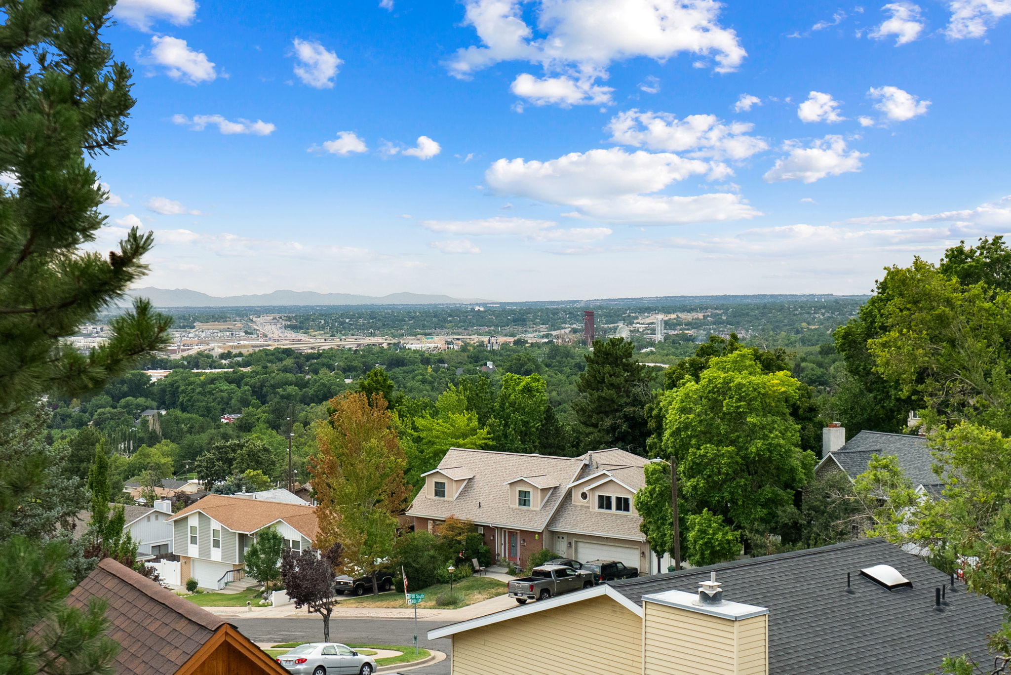 Elevated perspective showcasing a broad urban landscape framed by mature trees, with distant mountains under a blue sky with clouds